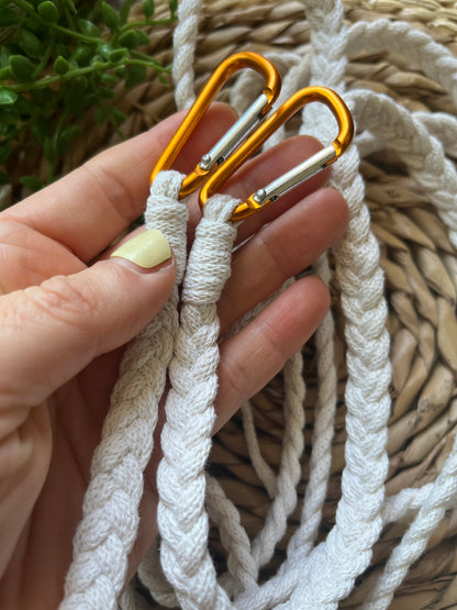 A hand holding orange carabiner clips on the end of a white braided apron strap.