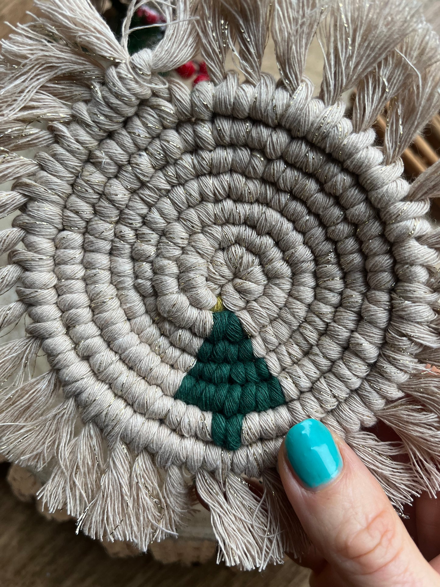 A hand holding a round macrame coaster with a small christmas tree accent and fringed edge. Handmade with recycled cotton yarn in a sparkly gold colour. 