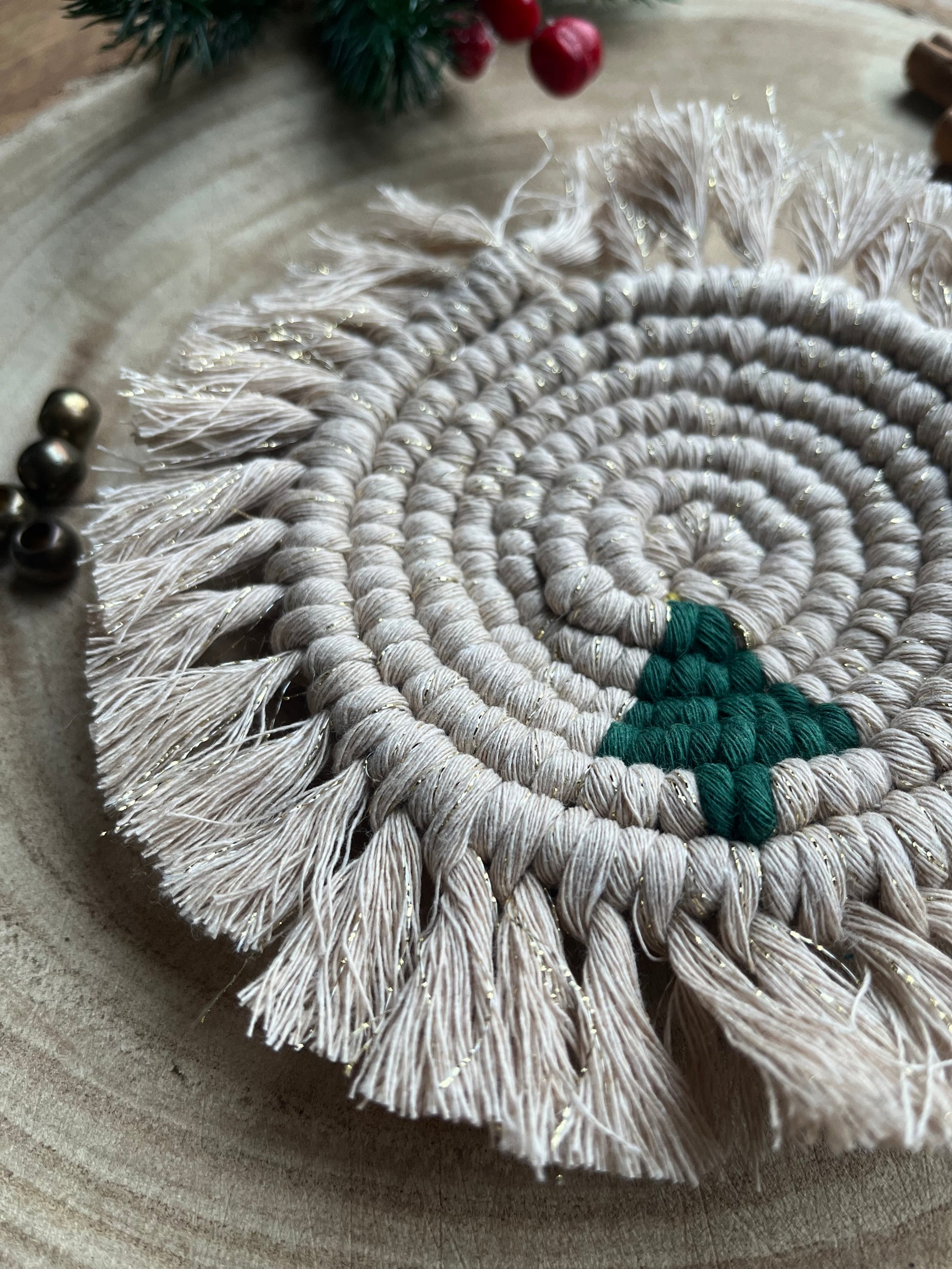 Close up image of a round macrame coaster with a small christmas tree accent and fringed edge. Handmade with recycled cotton yarn in sparkly gold. 