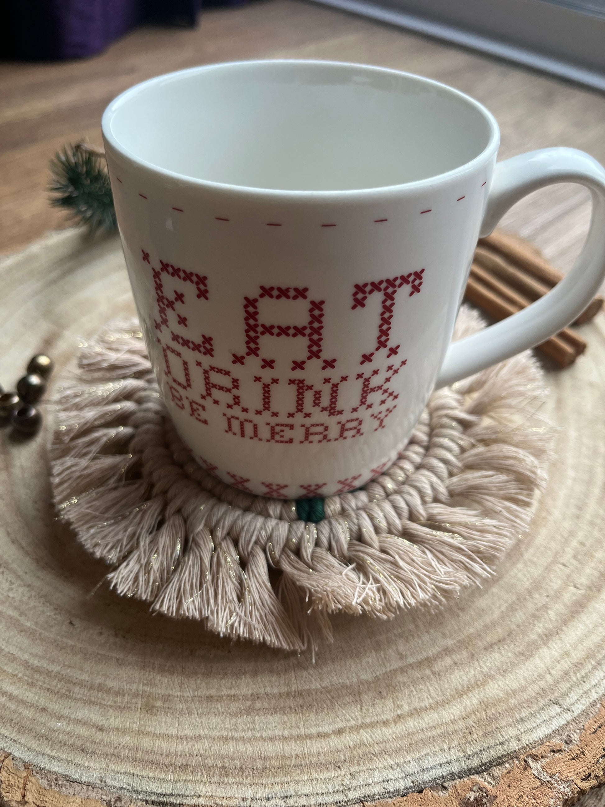 A white mug with red text sits on a round macrame coaster with a fringed edge. Handmade with recycled cotton yarn in a gold sparkly colour. 