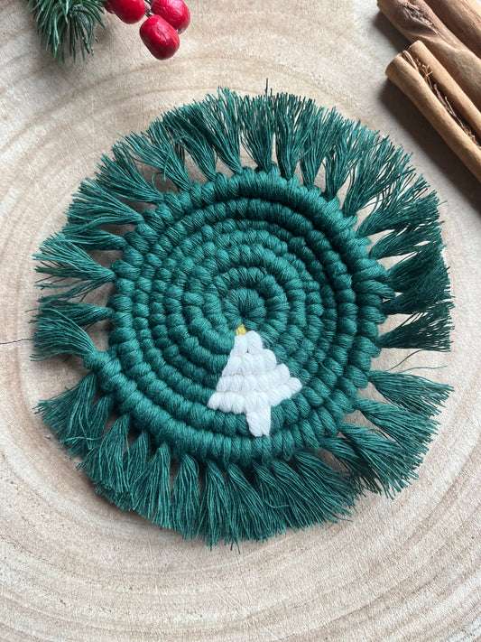 A handmade macrame round coaster with a Christmas Tree detail in the centre, and fringed edge, displayed on a wooden surface.