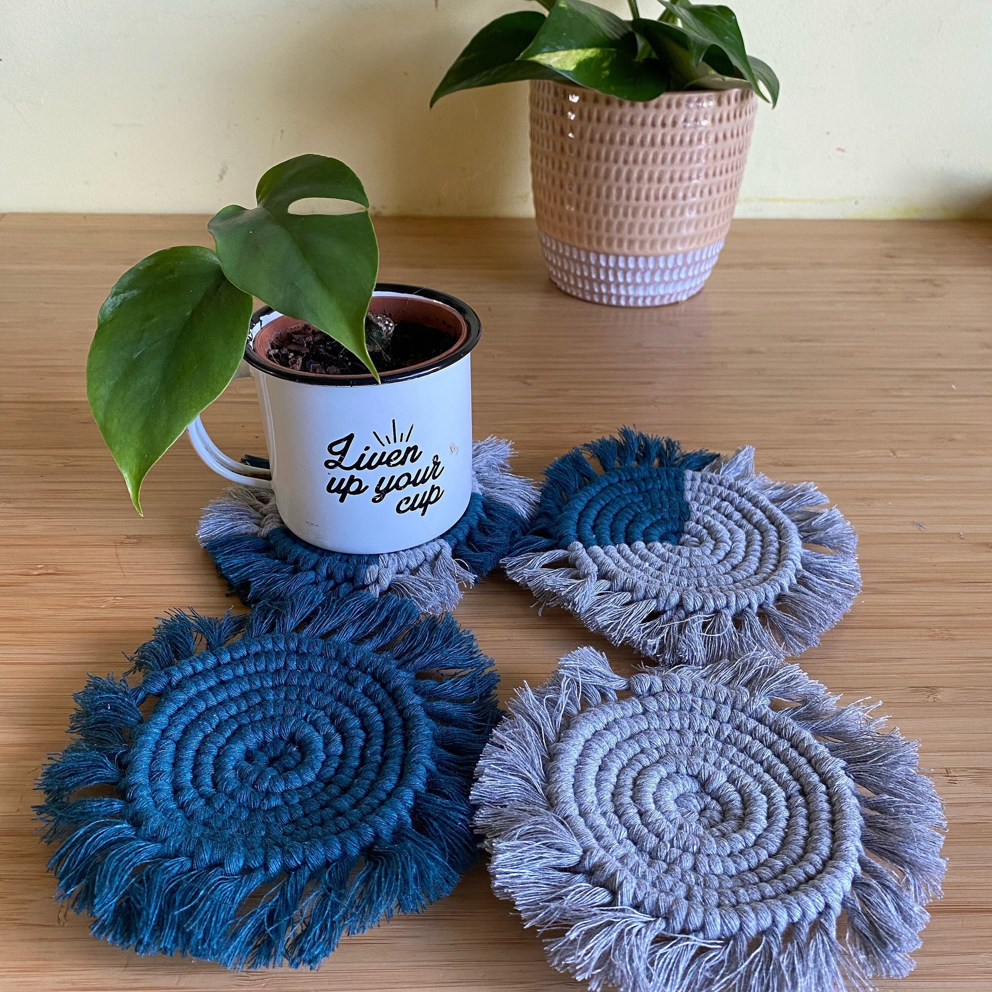 A set of 4 round macrame coasters made with blush pink and natural yarns, with a soft fringed edge, are placed on a wooden table, with a small green houseplant in a metal mug placed on top of one of the coasters. There is a larger green houseplant in a ceramic plant pot on the table in the background. 