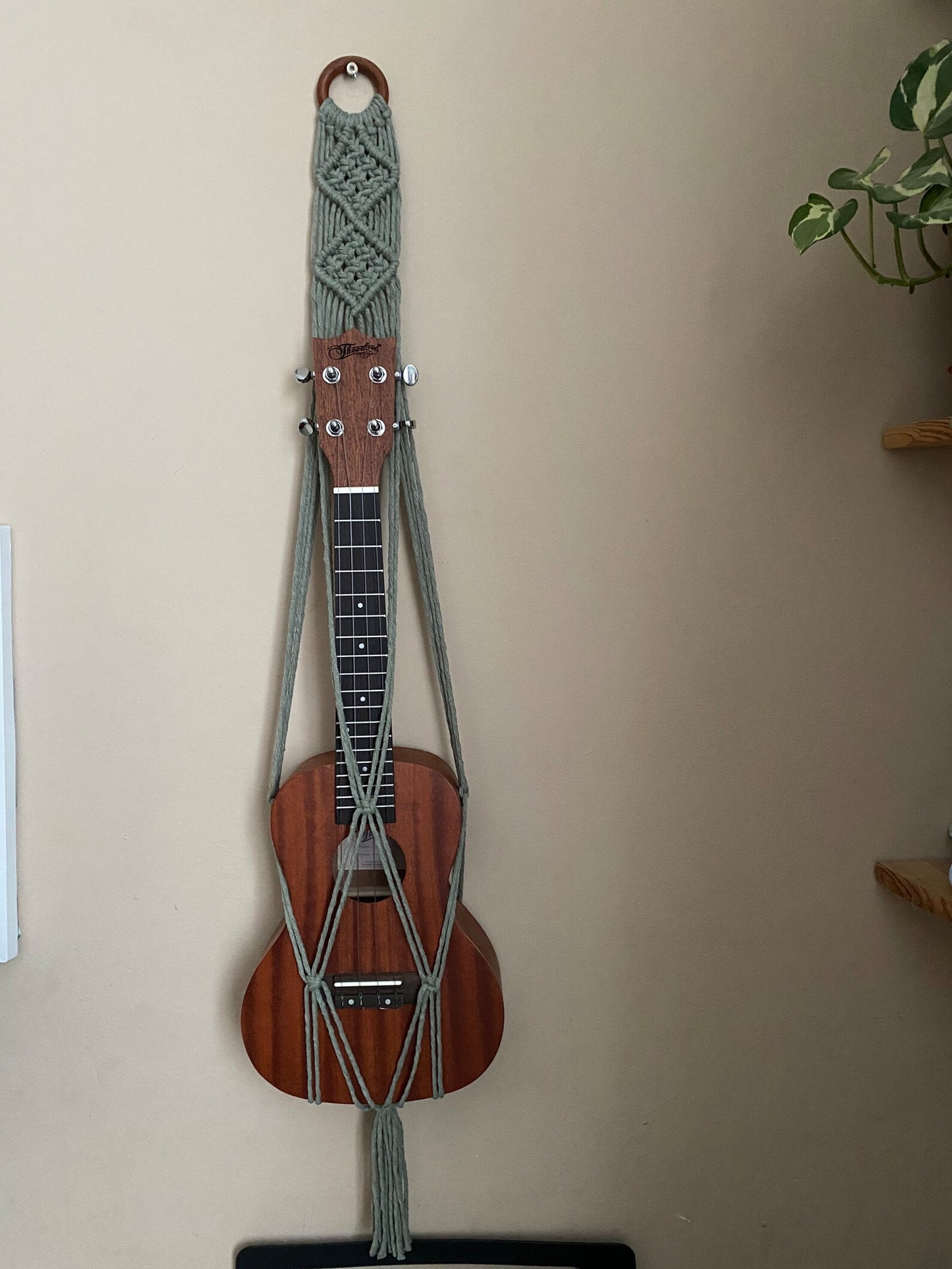 A dark brown ukulele sits inside of a handmade macrame wall hanging ukulele hanger, which is made with sage green recycled cotton, and a repurposed wooden hanging ring. 