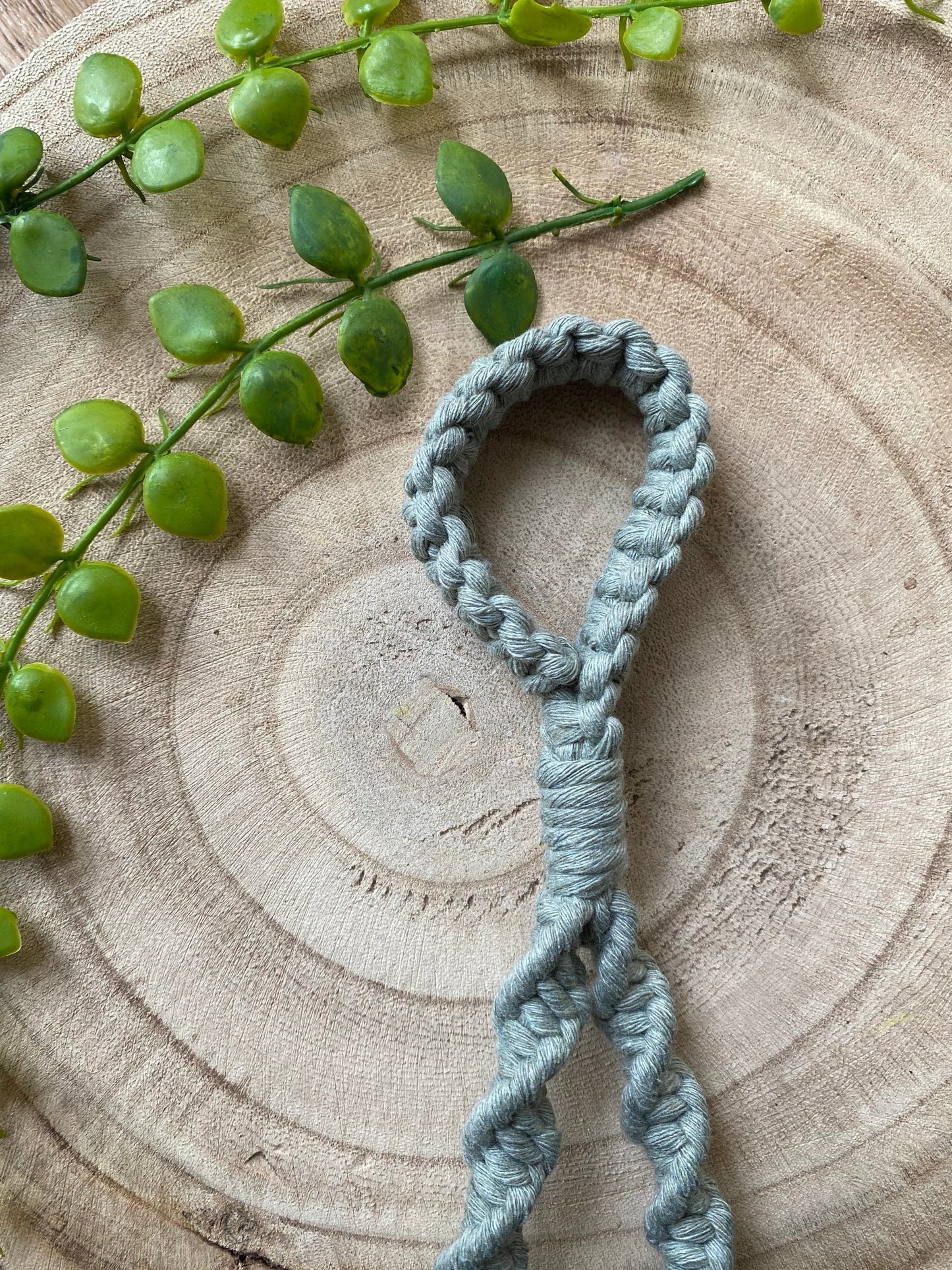 A coiled macrame loop made of light gray cord rests on a round wooden surface with a circular pattern. Green string beads are placed along the top-left edge, adding a natural element to this eco-friendly decor. This piece doubles as the Macra-Made-With-Love Single Toilet Roll Holder, perfect for any bathroom accessory collection.