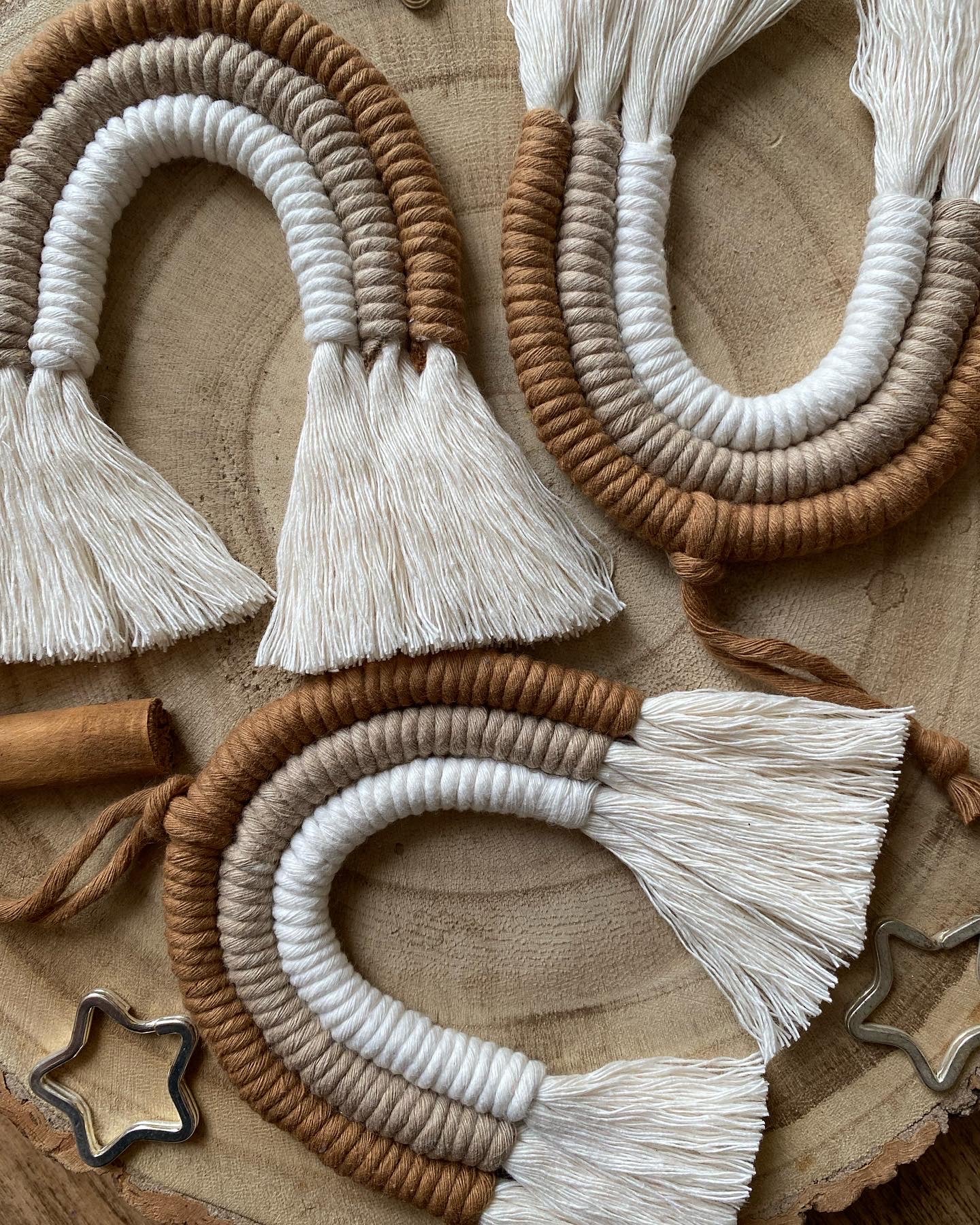 A set of three handmade macrame rainbow Christmas decorations in brown, white and beige colours with white tassels, displayed on a wooden surface.