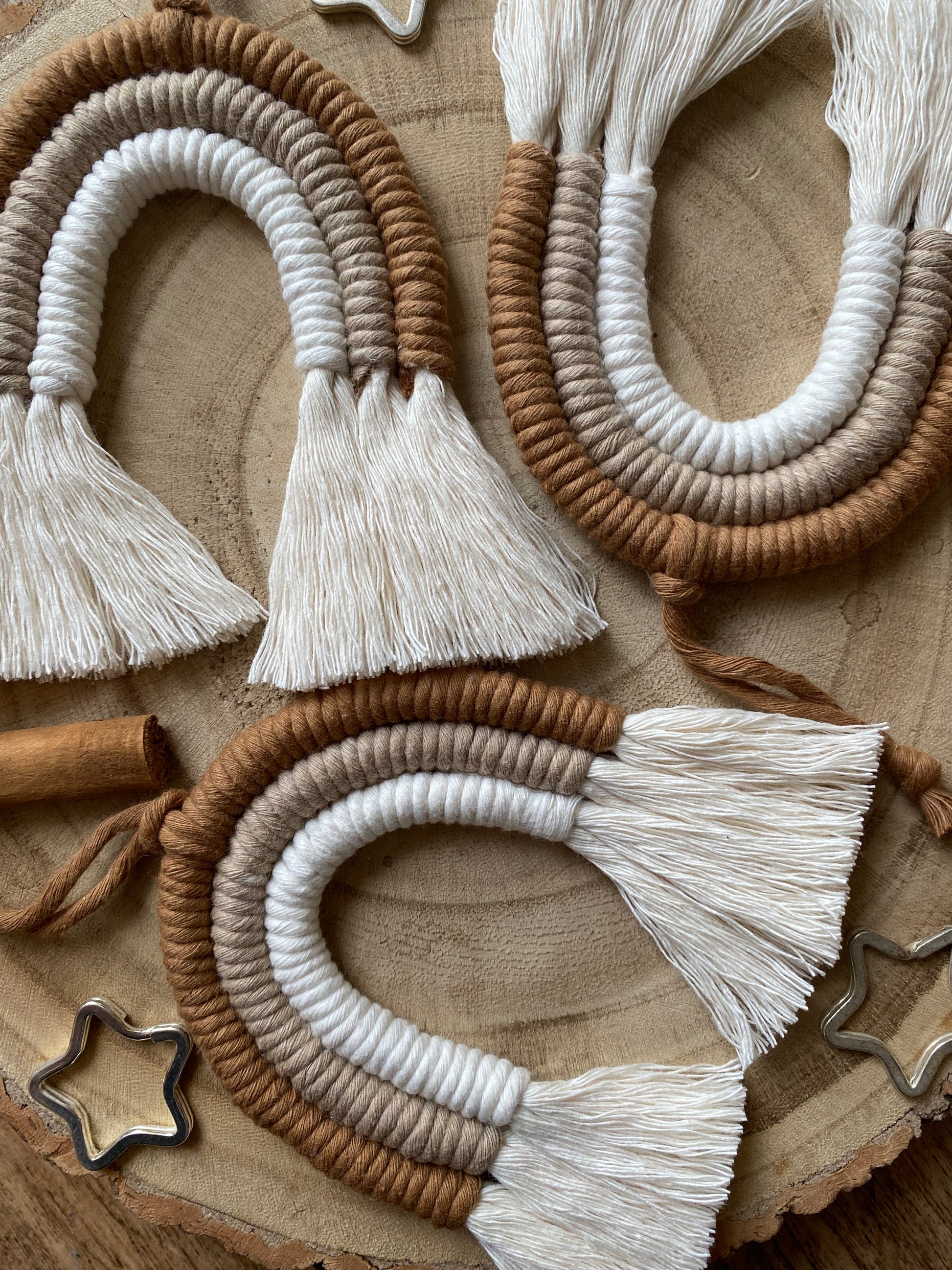 Close up of a set of three handmade macrame rainbow Christmas decorations in brown, beige and white yarn with white tassels, displayed on a wooden surface with beads and cinnamon sticks for decoration. 