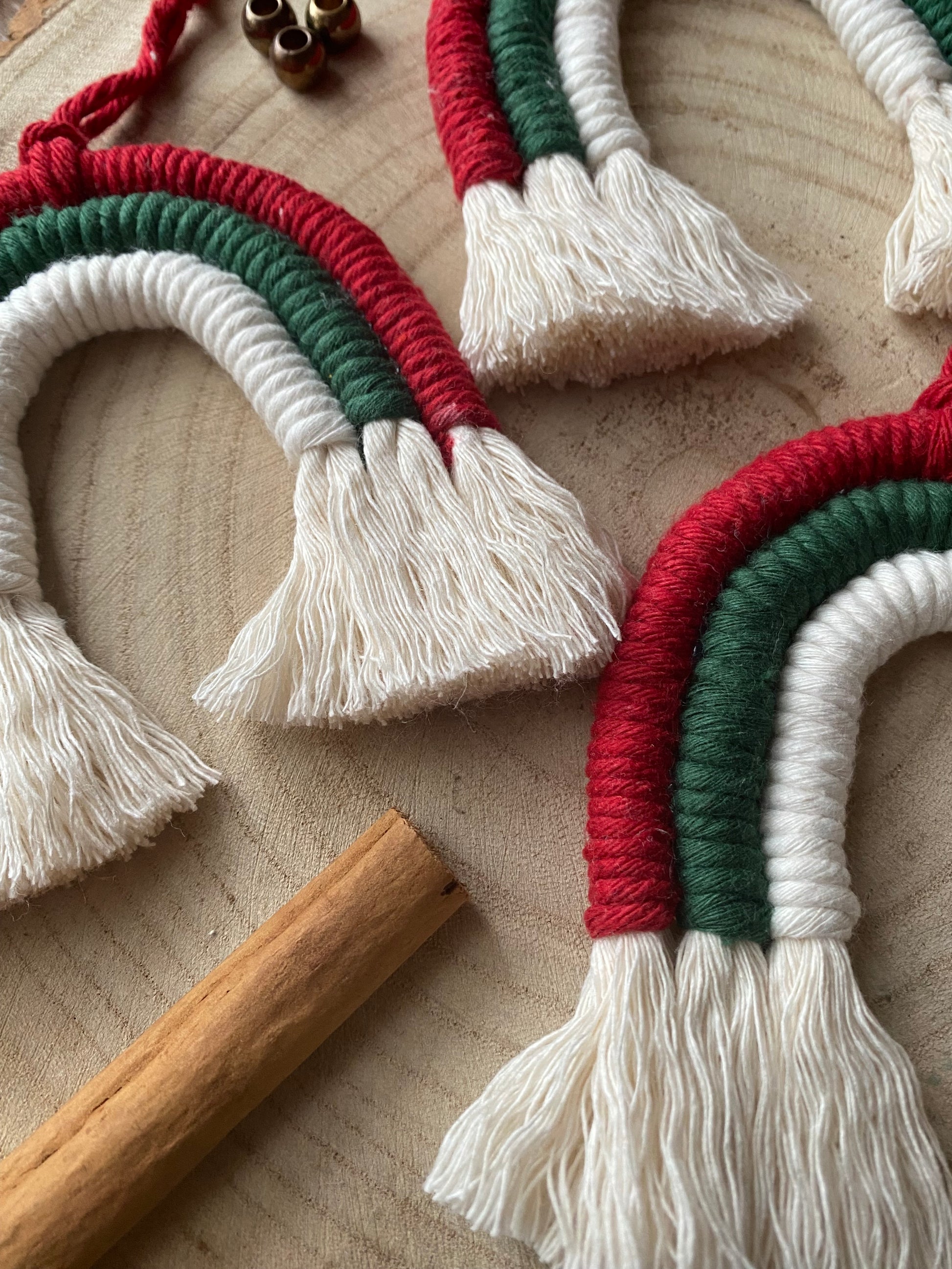 Close up of a set of three handmade macrame rainbow Christmas decorations in red, green, and white yarn with white tassels, displayed on a wooden surface with beads and cinnamon sticks for decoration. 