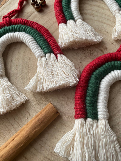 Close up of a set of three handmade macrame rainbow Christmas decorations in red, green, and white yarn with white tassels, displayed on a wooden surface with beads and cinnamon sticks for decoration. 
