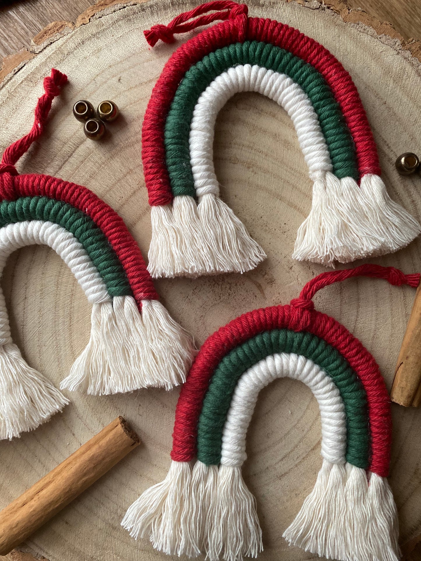 Close up of a set of three handmade macrame rainbow Christmas decorations in red, green, and white yarn with white tassels, displayed on a wooden surface with beads and cinnamon sticks for decoration. 