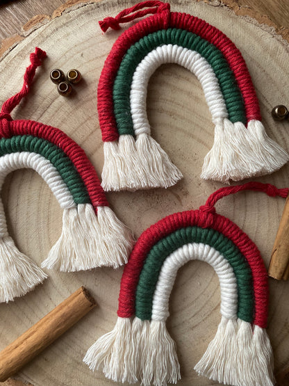 Close up of a set of three handmade macrame rainbow Christmas decorations in red, green, and white yarn with white tassels, displayed on a wooden surface with beads and cinnamon sticks for decoration. 