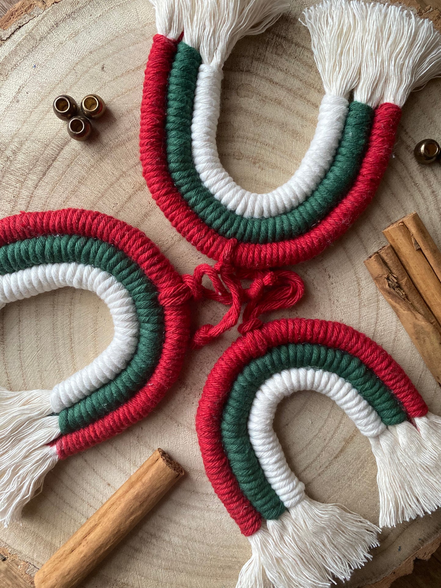 A set of three handmade macrame rainbow Christmas decorations in red, green, and white colours with white tassels, displayed on a wooden surface.