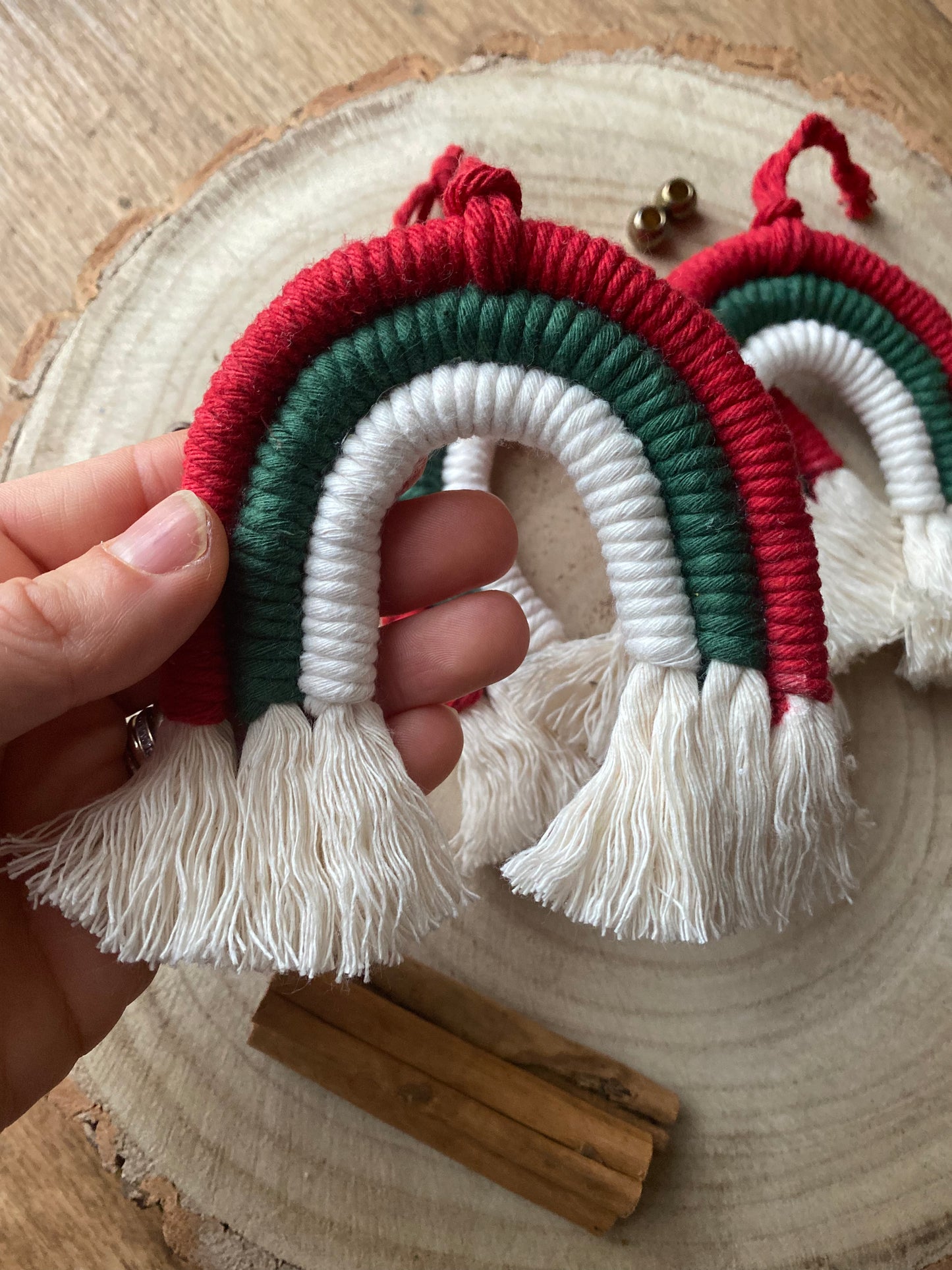 A hand holding one of a set of three handmade macrame rainbow Christmas decorations in red, green, and white yarn with white tassels, displayed on a wooden surface with cinnamon sticks for decoration. 