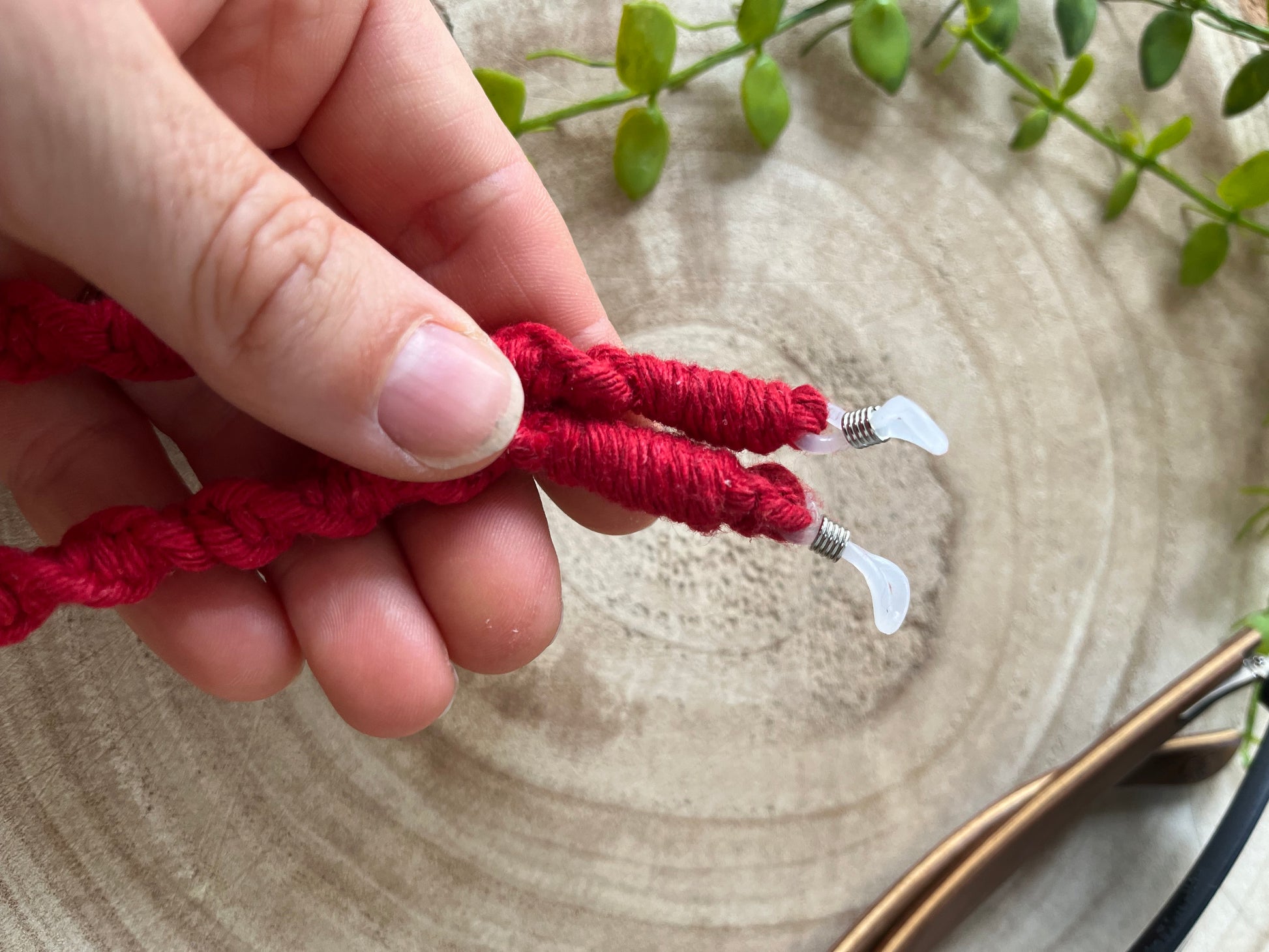 A hand holding a handmade macrame strap for glasses, made with red recycled cotton with clear loops on each end. 