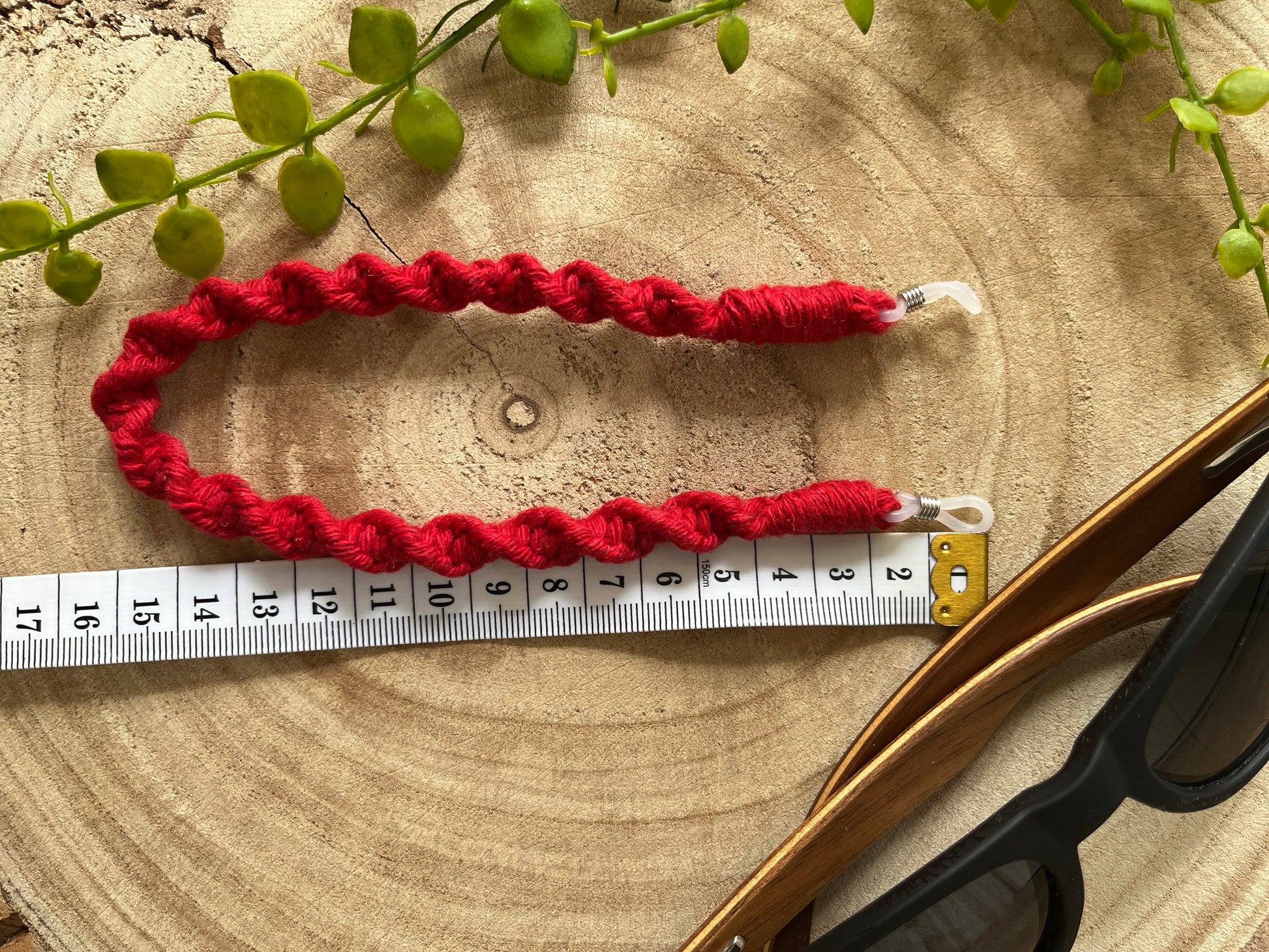 Handmade macrame short strap for glasses, made with red recycled cotton, laid on a wooden surface with a white measuring tape next to it. 