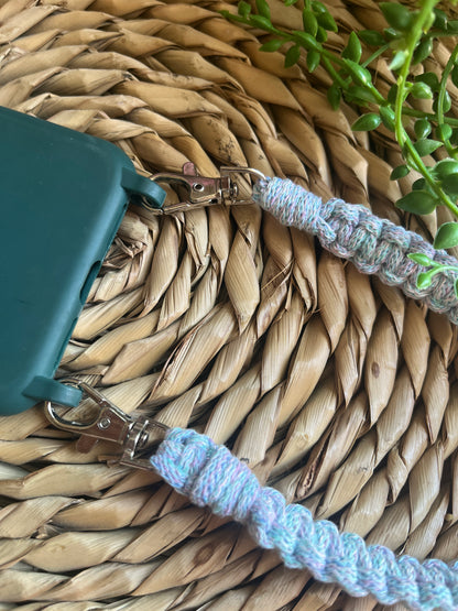 Blue braided wrist strap attached to a green phone case on a woven mat with a plant in the corner.
