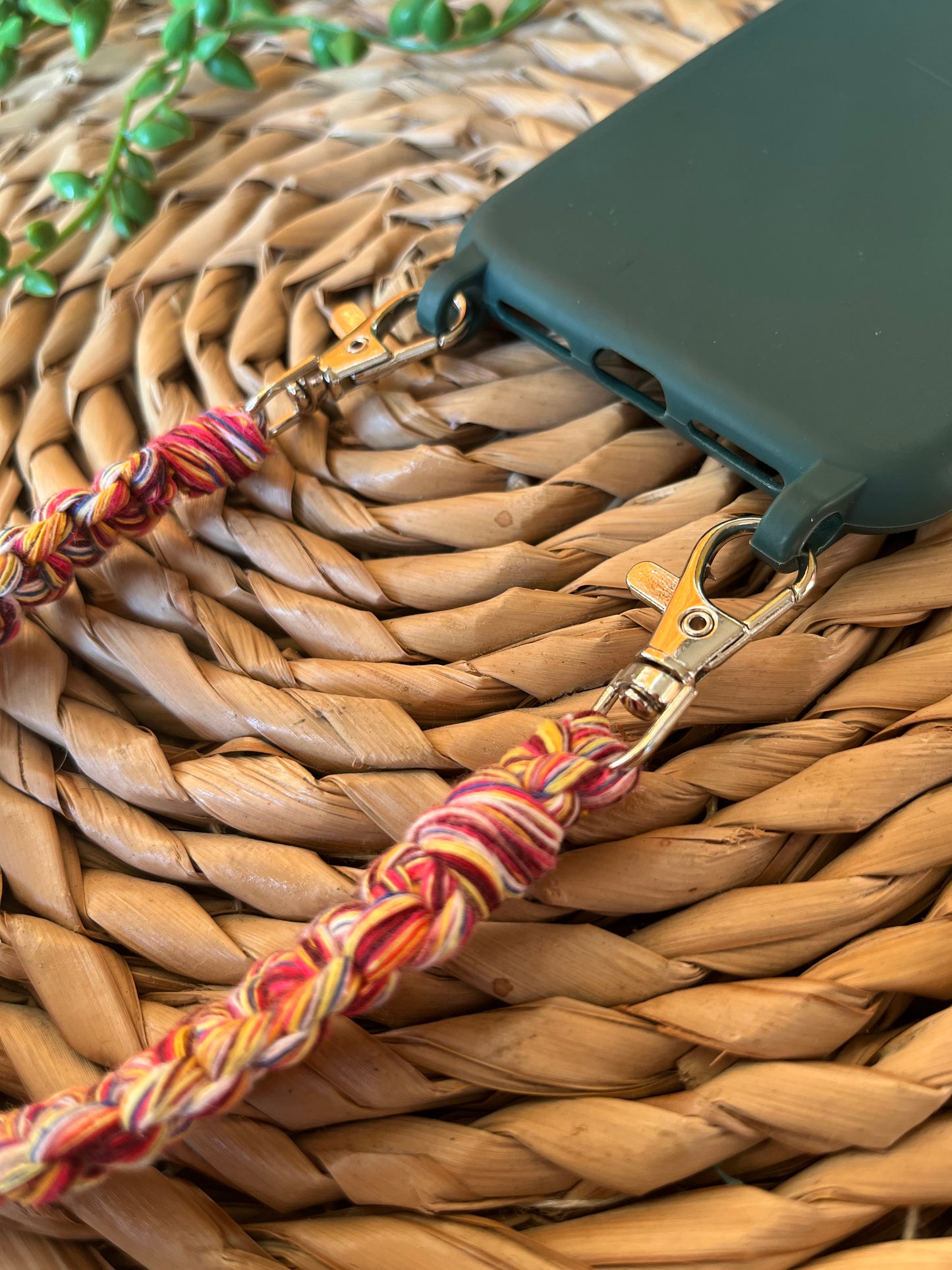 Handmade short macrame phone wristlet loop for phone case. Shown attached to a green phone case on a wicker mat. Handmade with multicoloured recycled cotton yarn, with a small metal lobster clasp on each end. 