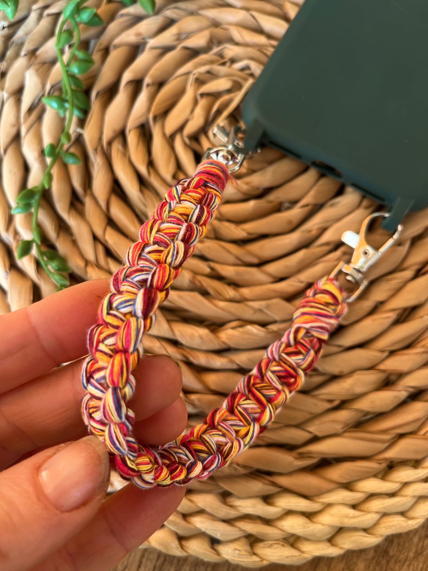 A hand holding a handmade short macrame phone wristlet loop for phone case. Shown attached to a green phone case on a wicker mat. Handmade with multicoloured recycled cotton yarn, with a small metal lobster clasp on each end. 