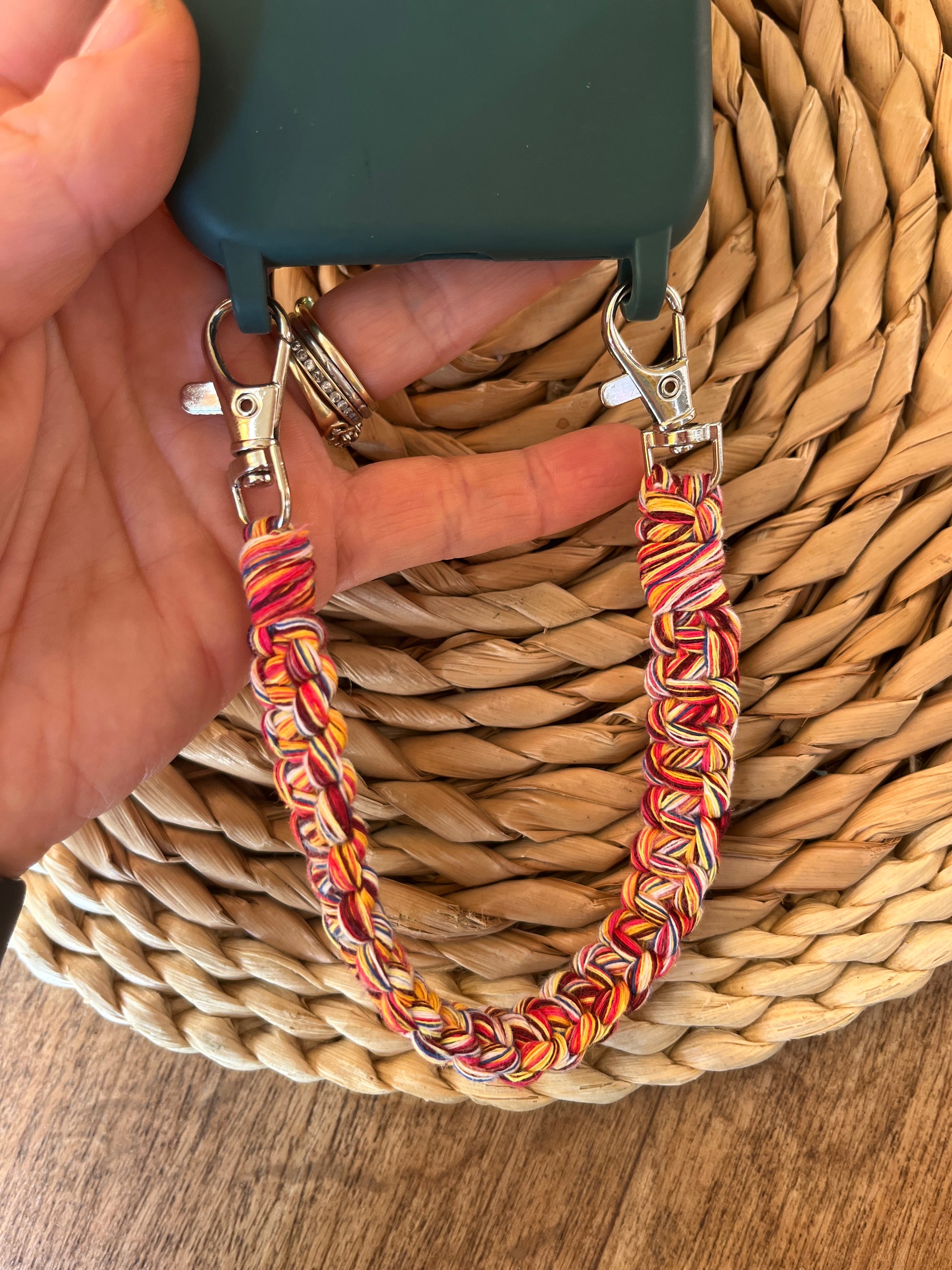 A hand holding a handmade short macrame phone wristlet loop for phone case. Shown attached to a green phone case on a wicker mat. Handmade with multicoloured recycled cotton yarn, with a small metal lobster clasp on each end. 