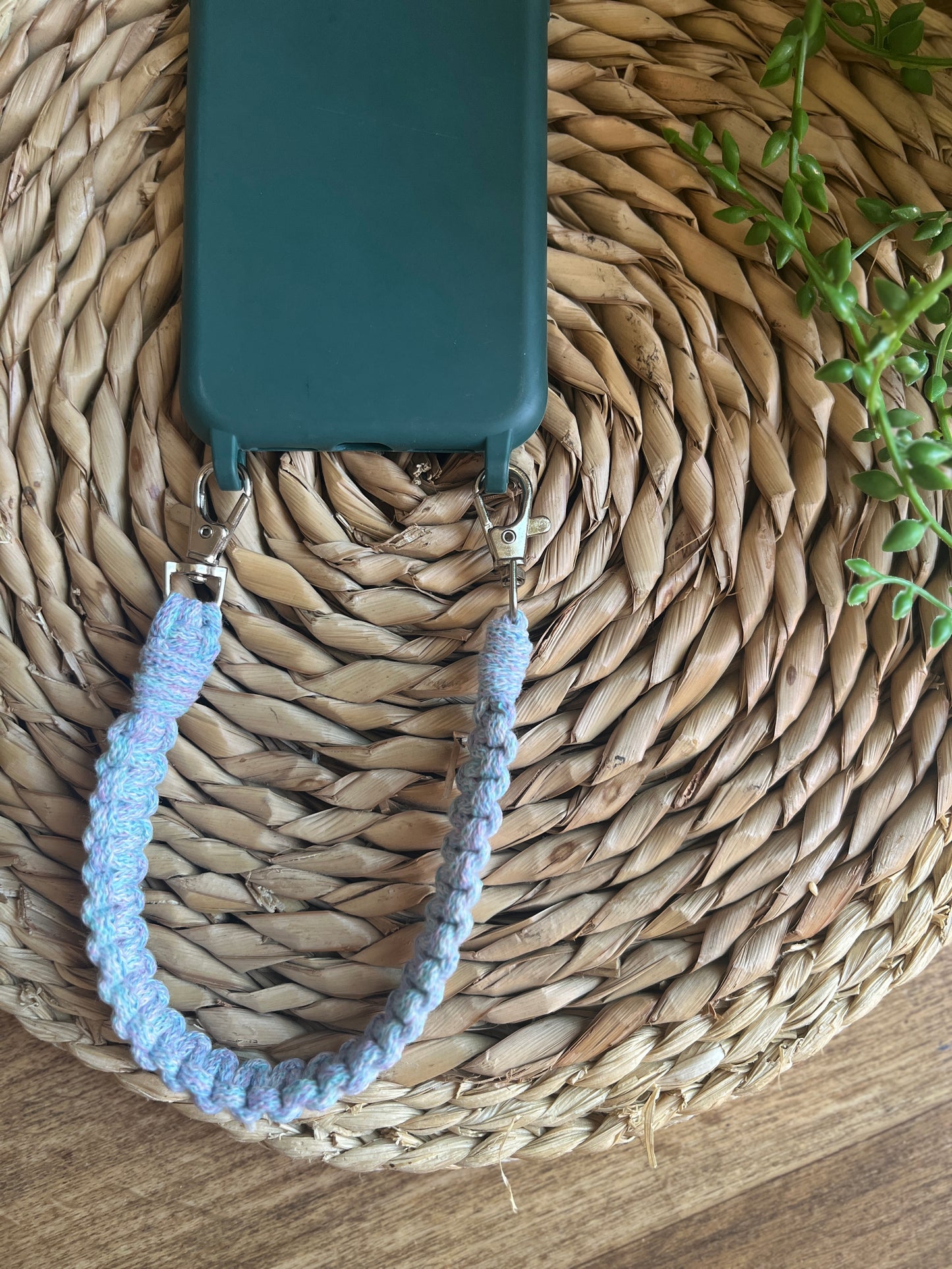 Handmade short macrame phone wristlet loop for phone case made with pastel yarn. Shown on a wicker mat, attached to the bottom of a green phone case. 