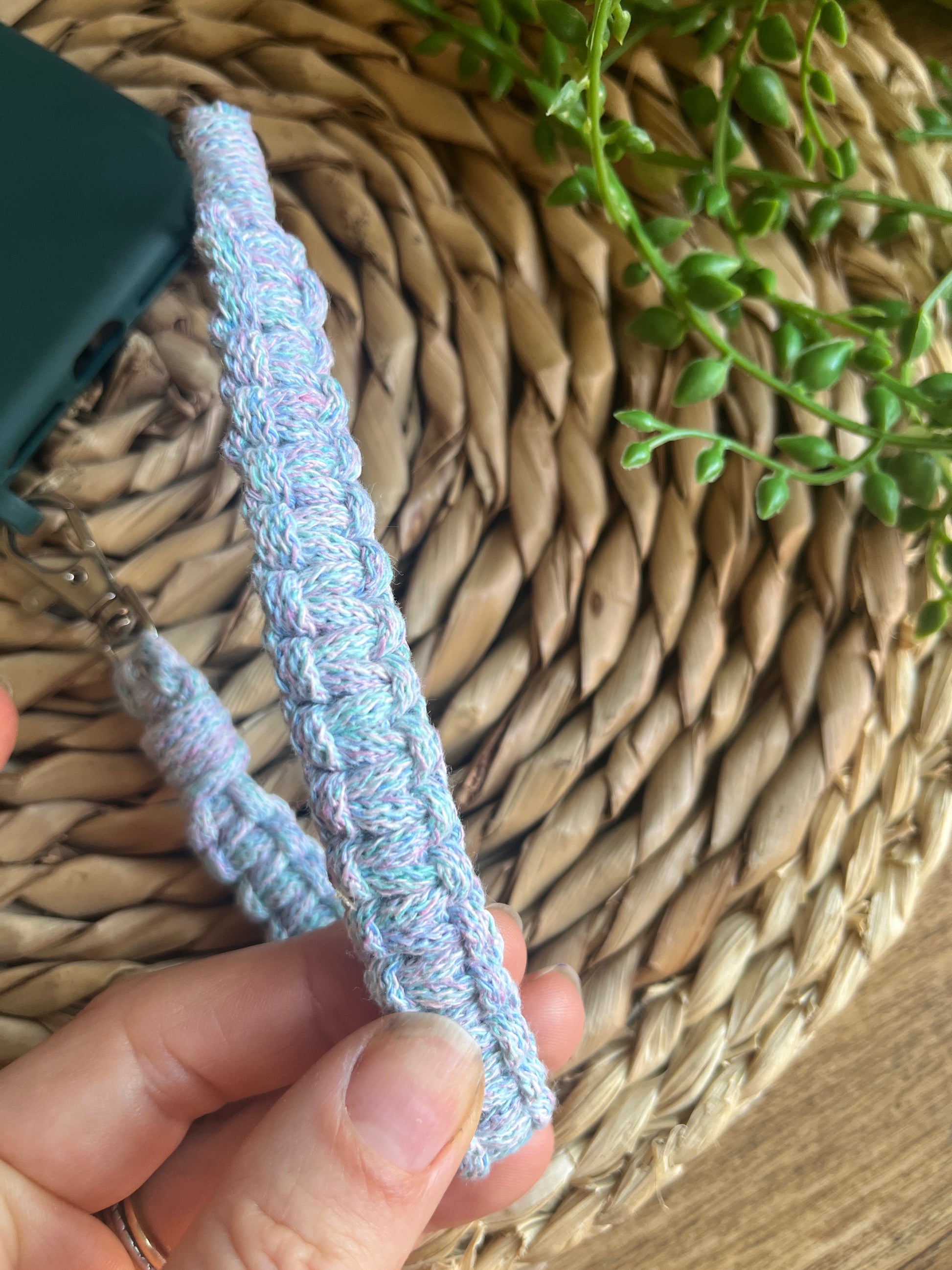 Hand holding a knotted wristlet on a woven mat with a plant in the corner.