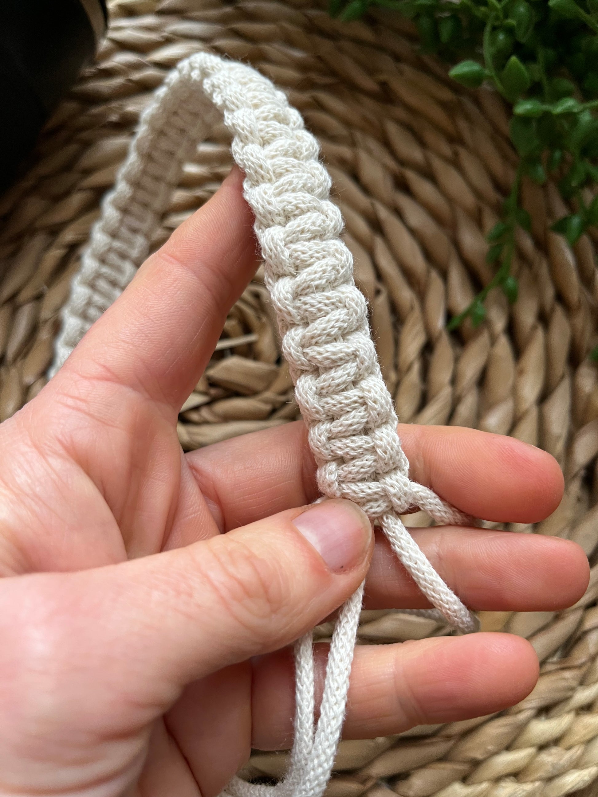 a hands holds a macrame coffee cup holder strap, it is made with natural recycled cotton and features a long section is neat knots. 