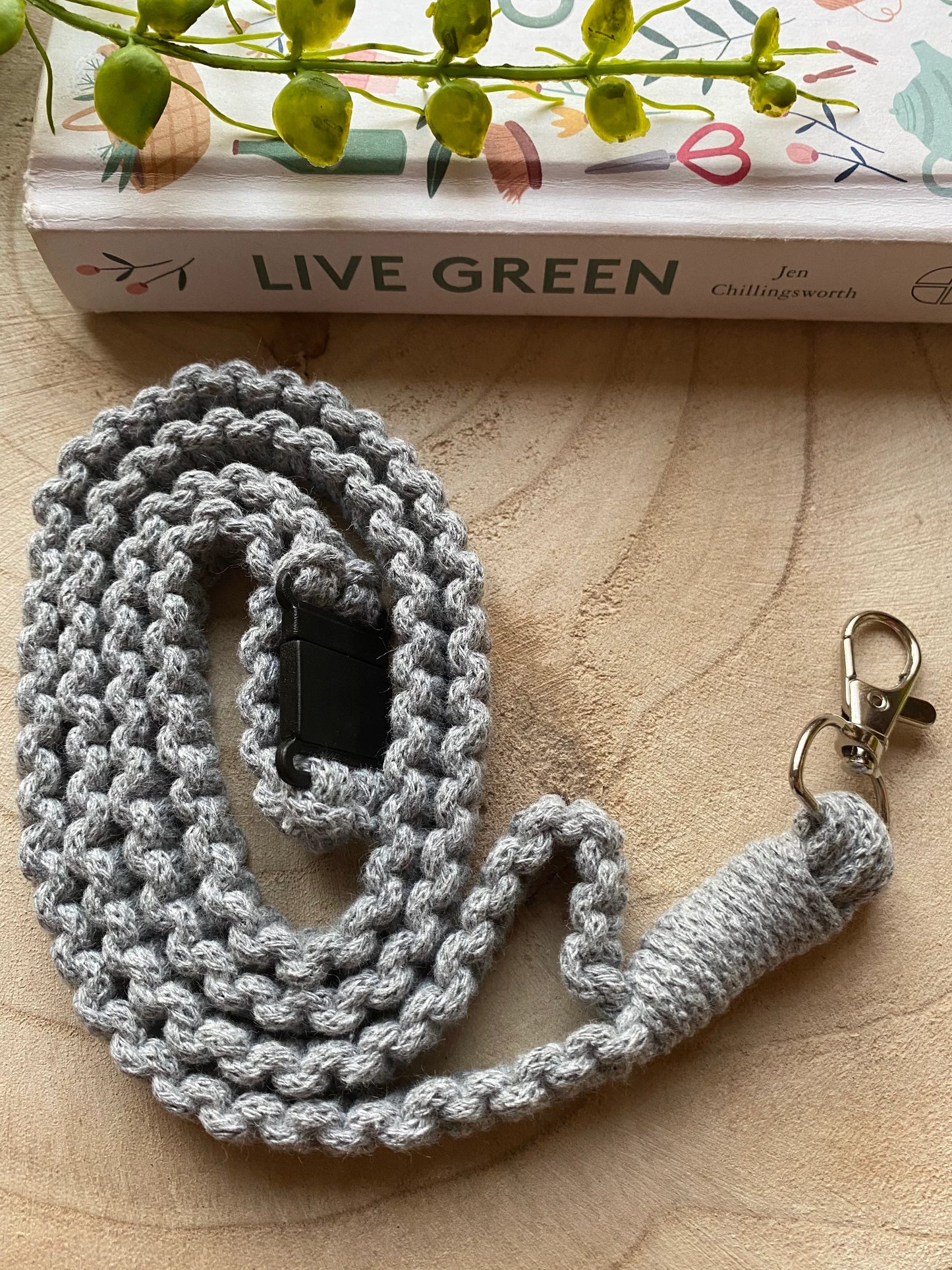 A coiled, gray, handmade Eco friendly lanyard badge holder - 34” by Macra-Made-With-Love with a metallic clasp and a plastic clip lies on a wooden surface. A book titled "Live Green" by Jen Chillingsworth, with an illustrated cover and a green plant garnish, are partially visible in the background. This eco-friendly lanyard also doubles as an ID badge holder.