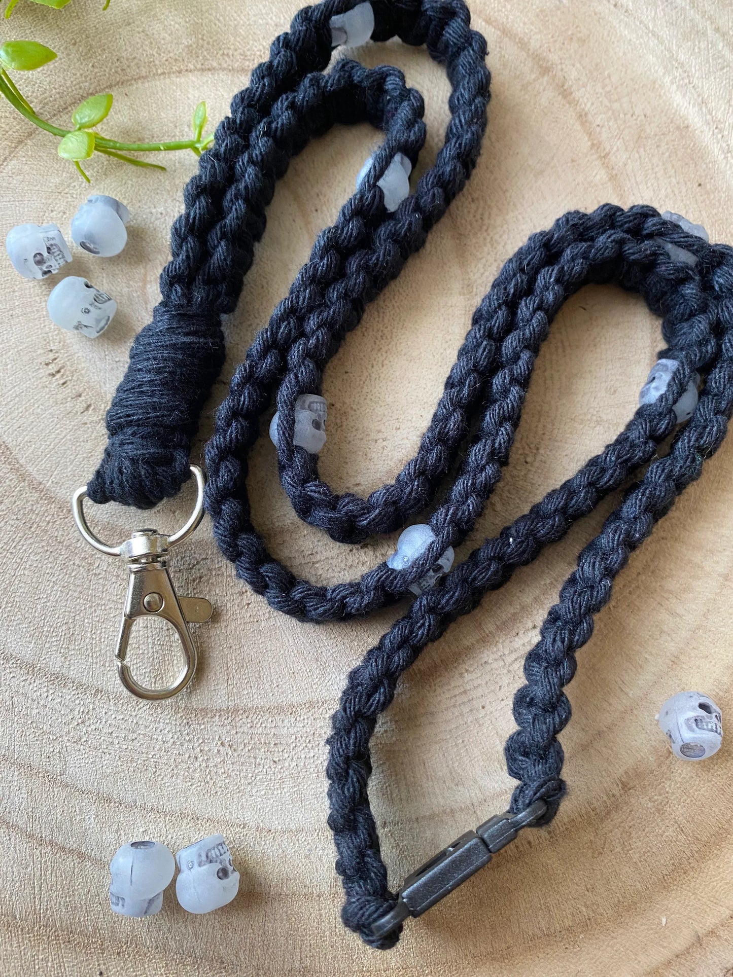 A close-up of the Macra-Made-With-Love Eco friendly lanyard badge holder - 34” with a metallic clasp, lying in a loose spiral on a wooden surface. Transparent skull-shaped beads are woven intermittently in the eco-friendly lanyard. Several small, similar skull beads are scattered around. A green plant is in the background.