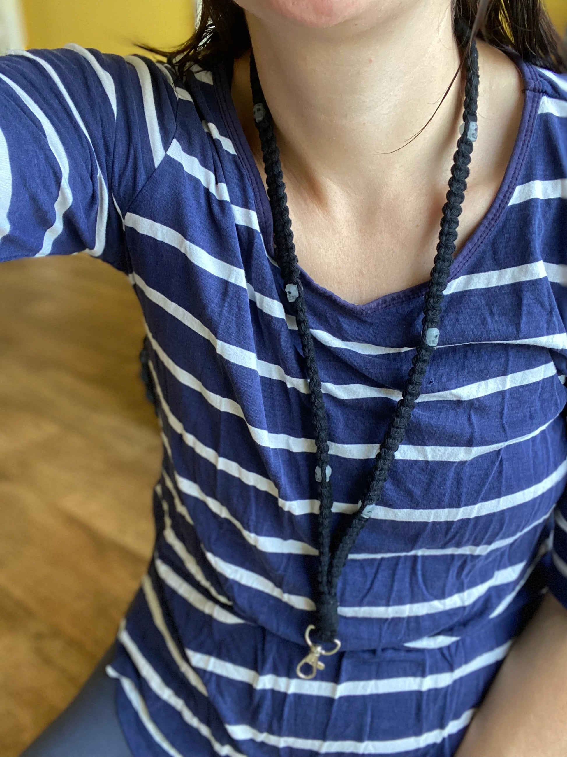 A person wearing a blue and white striped long-sleeve shirt and a long, dark-colored beaded necklace with a pendant takes a selfie. The background is indoors with part of a wooden floor visible. The Eco friendly lanyard badge holder - 34” from Macra-Made-With-Love adds an extra touch of uniqueness, though the person's face is partially out of the frame.