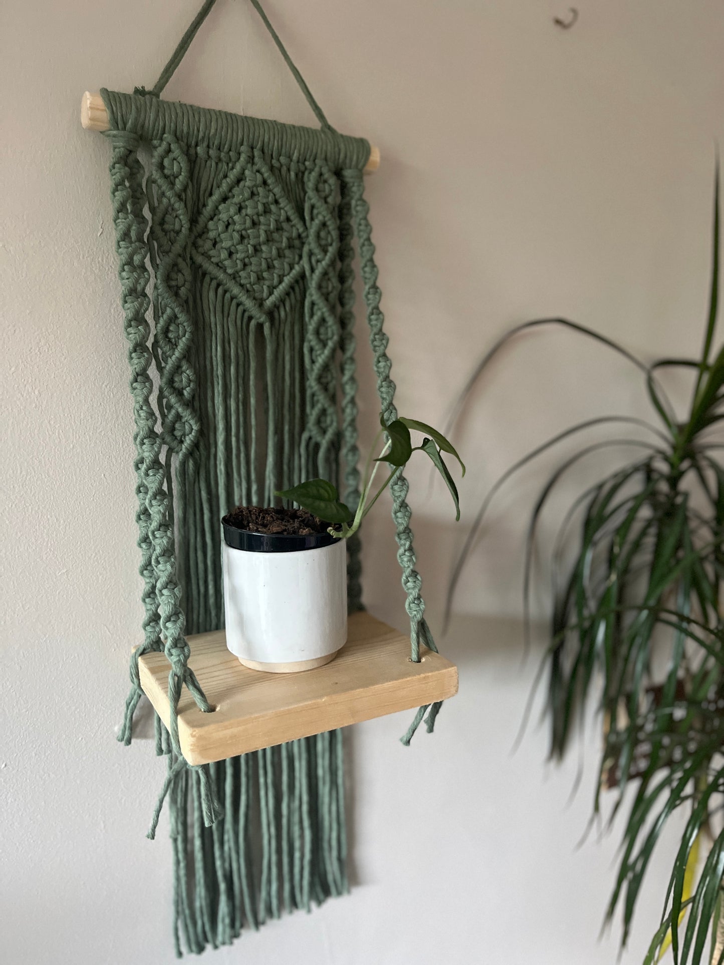 Macrame wall hanging with a wooden shelf holding a plant against a neutral wall.
