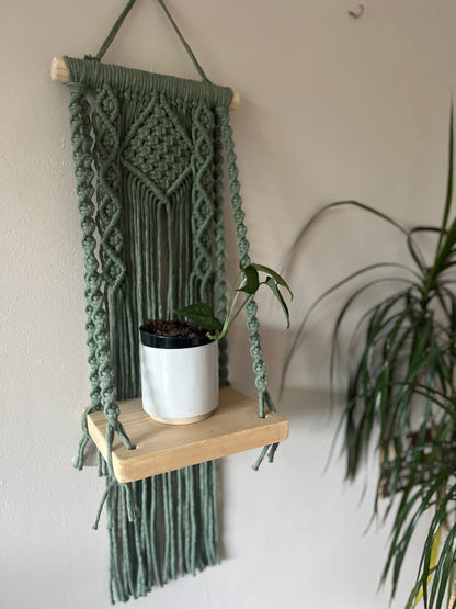 Macrame wall hanging with a wooden shelf holding a plant against a neutral wall.