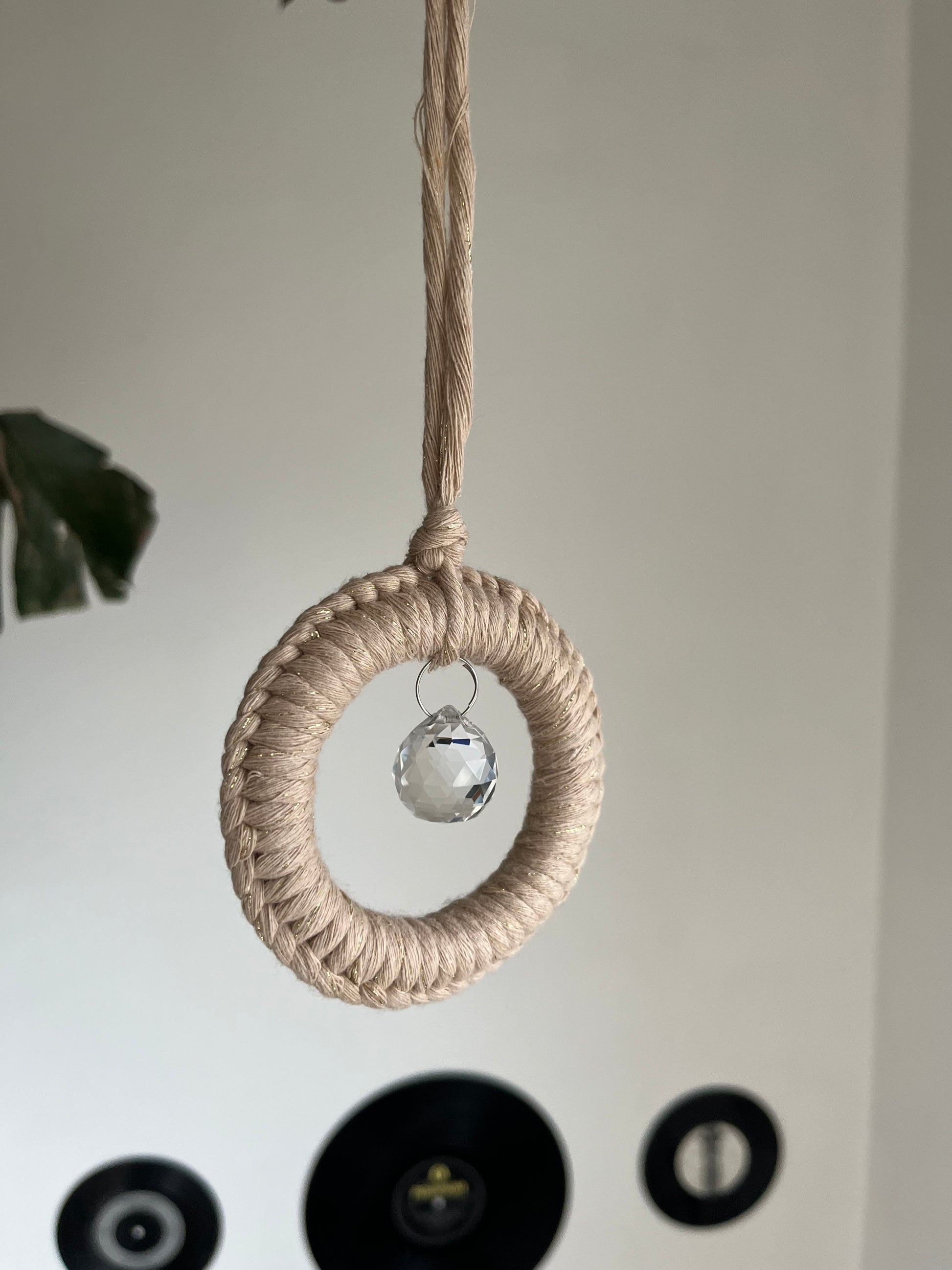 A handmade macrame circular hanging suncatcher, made with recycled cotton in sparkly gold with a small crystal prism. There are records hanging on a white wall in the background. 