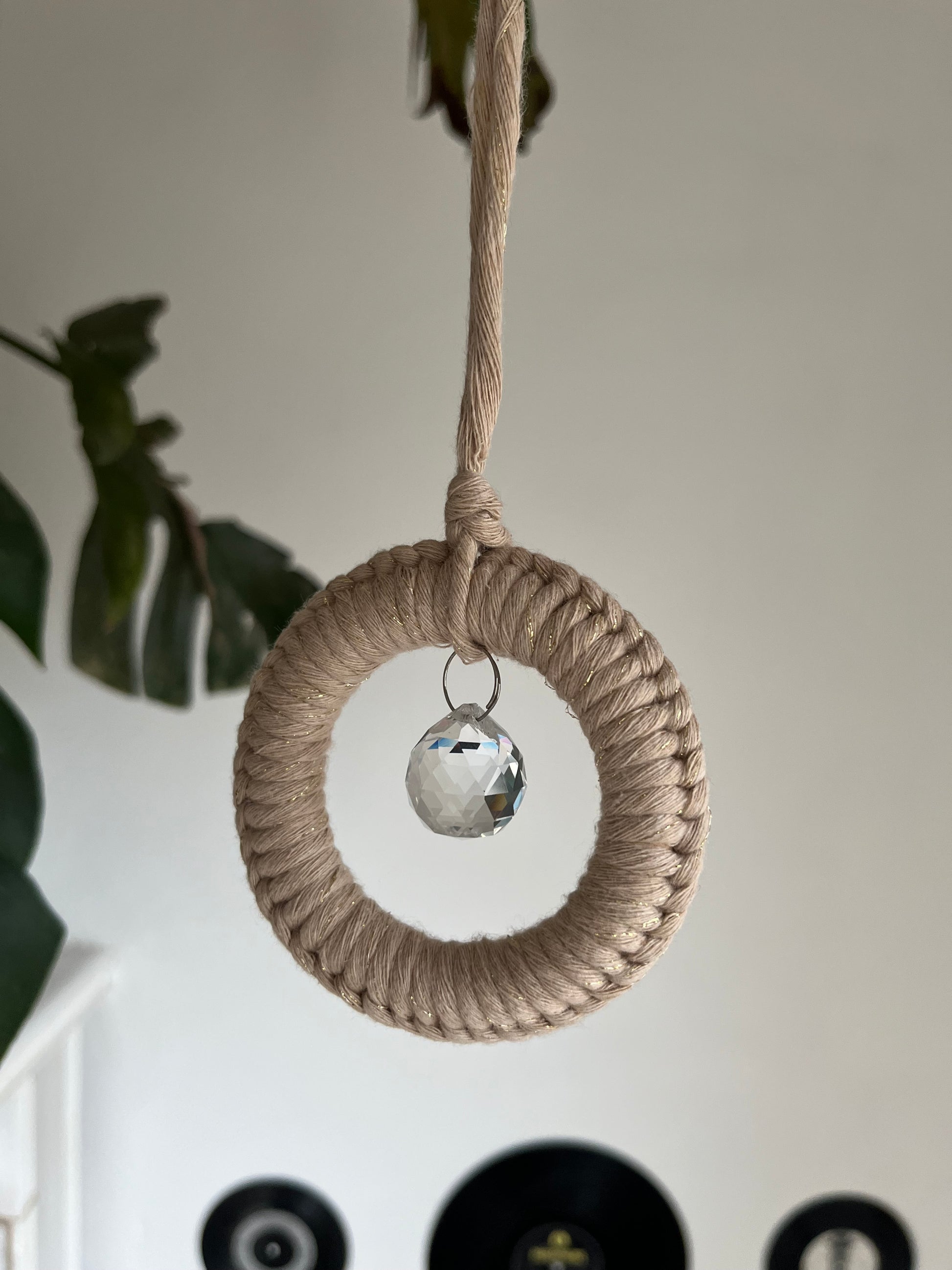 A handmade macrame circular hanging suncatcher, made with recycled cotton in sparkly gold with a small crystal prism. There are records hanging on a white wall in the background. 
