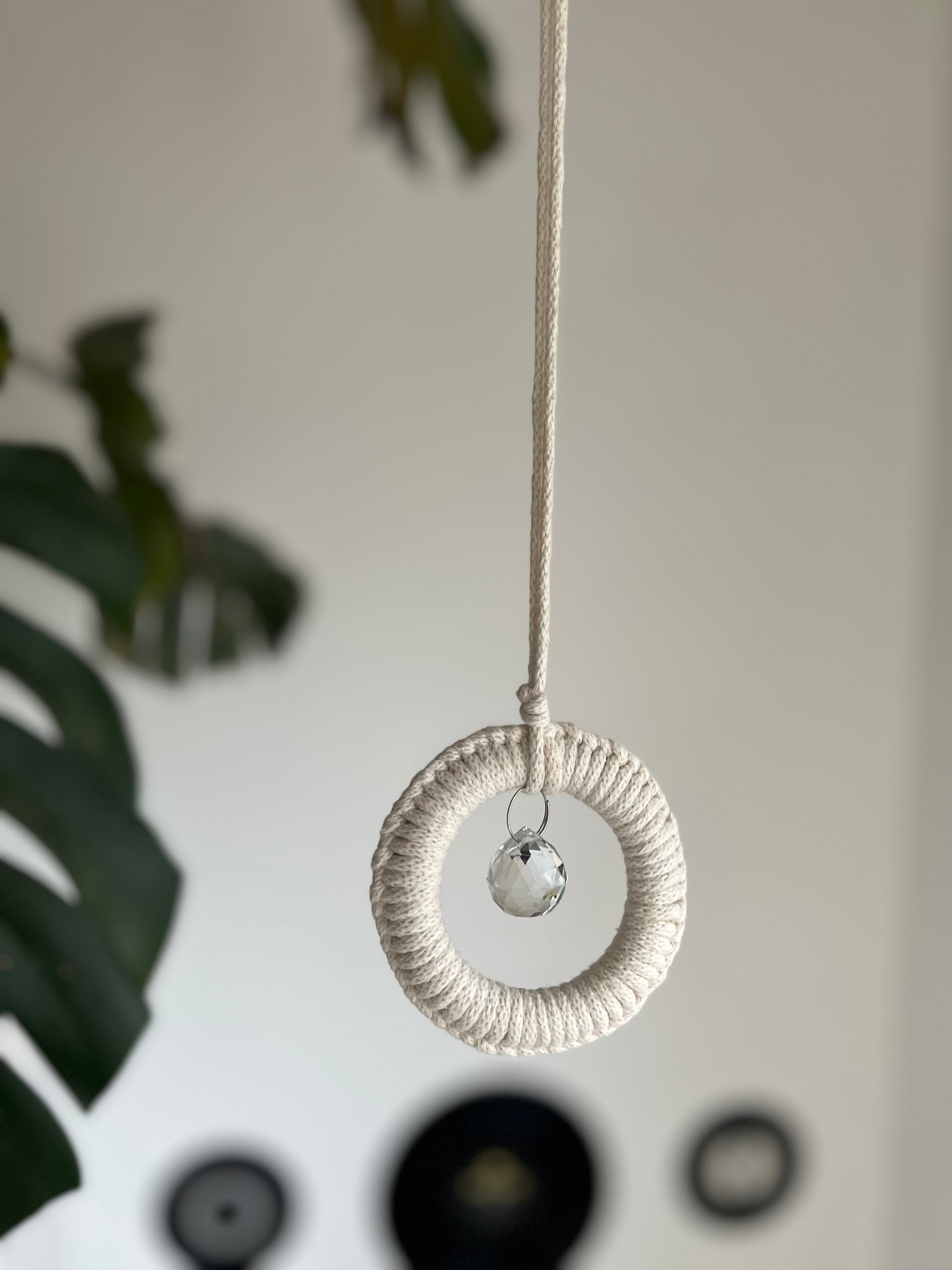 A handmade macrame circular hanging suncatcher, made with recycled cotton in an of white colour, with a small crystal prism. There is a green leafy plant in the background. 
