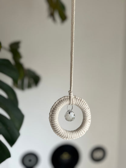 A handmade macrame circular hanging suncatcher, made with recycled cotton in an of white colour, with a small crystal prism. There is a green leafy plant in the background. 