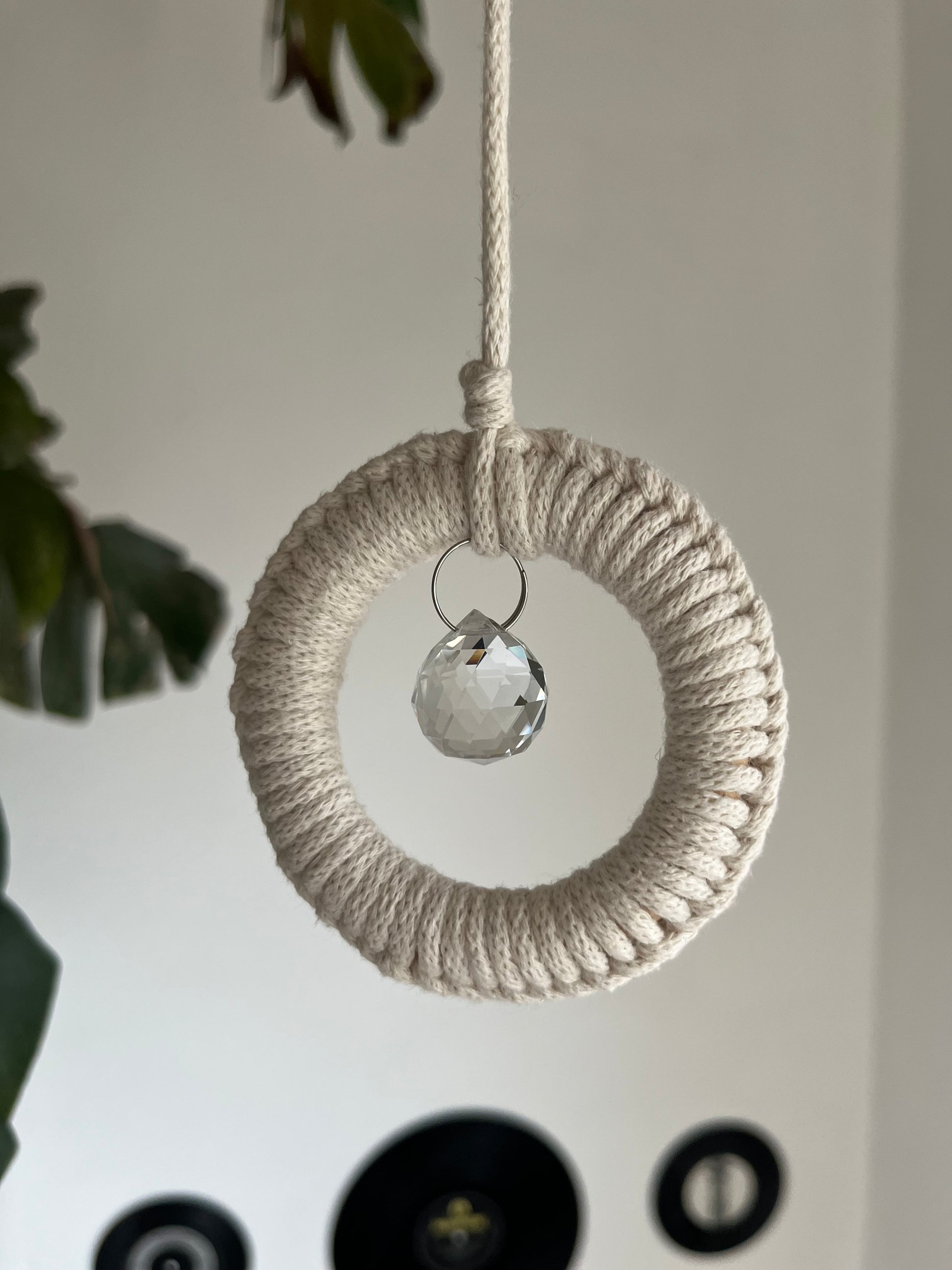 A handmade macrame circular hanging suncatcher, made with recycled cotton in an of white colour, with a small crystal prism. There is a green leafy plant in the background. 