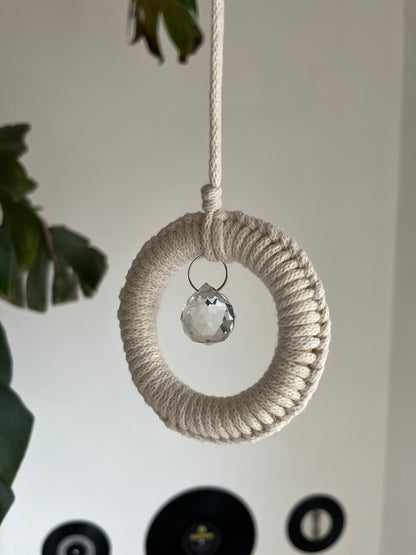 A handmade macrame circular hanging suncatcher, made with recycled cotton in an of white colour, with a small crystal prism. There is a green leafy plant in the background. 
