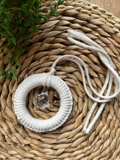 A handmade macrame circular hanging suncatcher, made with recycled cotton in an off white natural colour with a small crystal prism. The suncatcher is laid on a round wicker surface. 