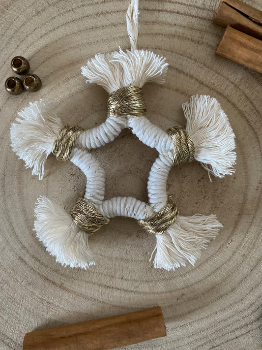 A small macrame star shaped hanging decoration, made with natural yarn with gold detailing, and fringed ends, laying on a wooden board. There are cinnamon sticks and metal stars laid out nearby for decoration. 
