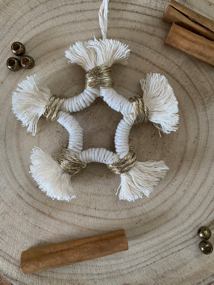 A small macrame star shaped hanging decoration, made with natural yarn with gold detailing, and fringed ends, laying on a wooden board. There are cinnamon sticks and metal stars laid out nearby for decoration. 
