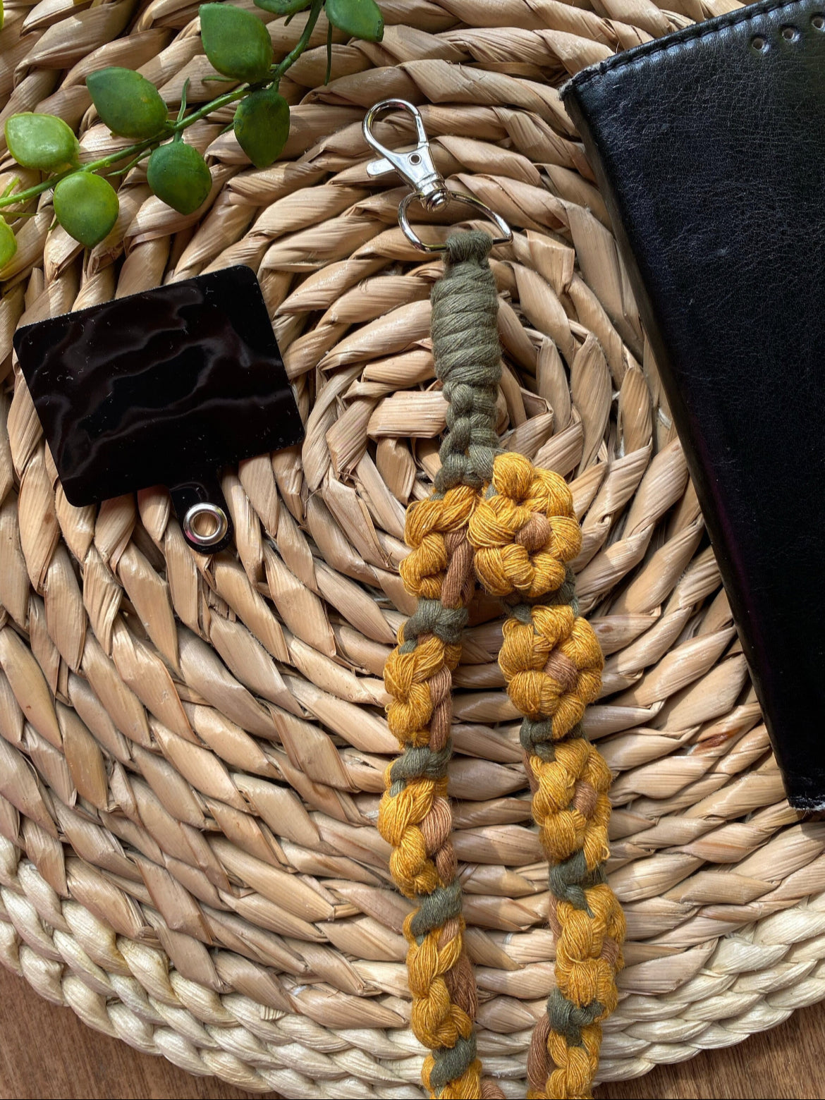 A black leather phone case placed on a woven mat, with a yellow and green sunflower-themed macramé phone charm wristlet and universal phone tether next to it. 