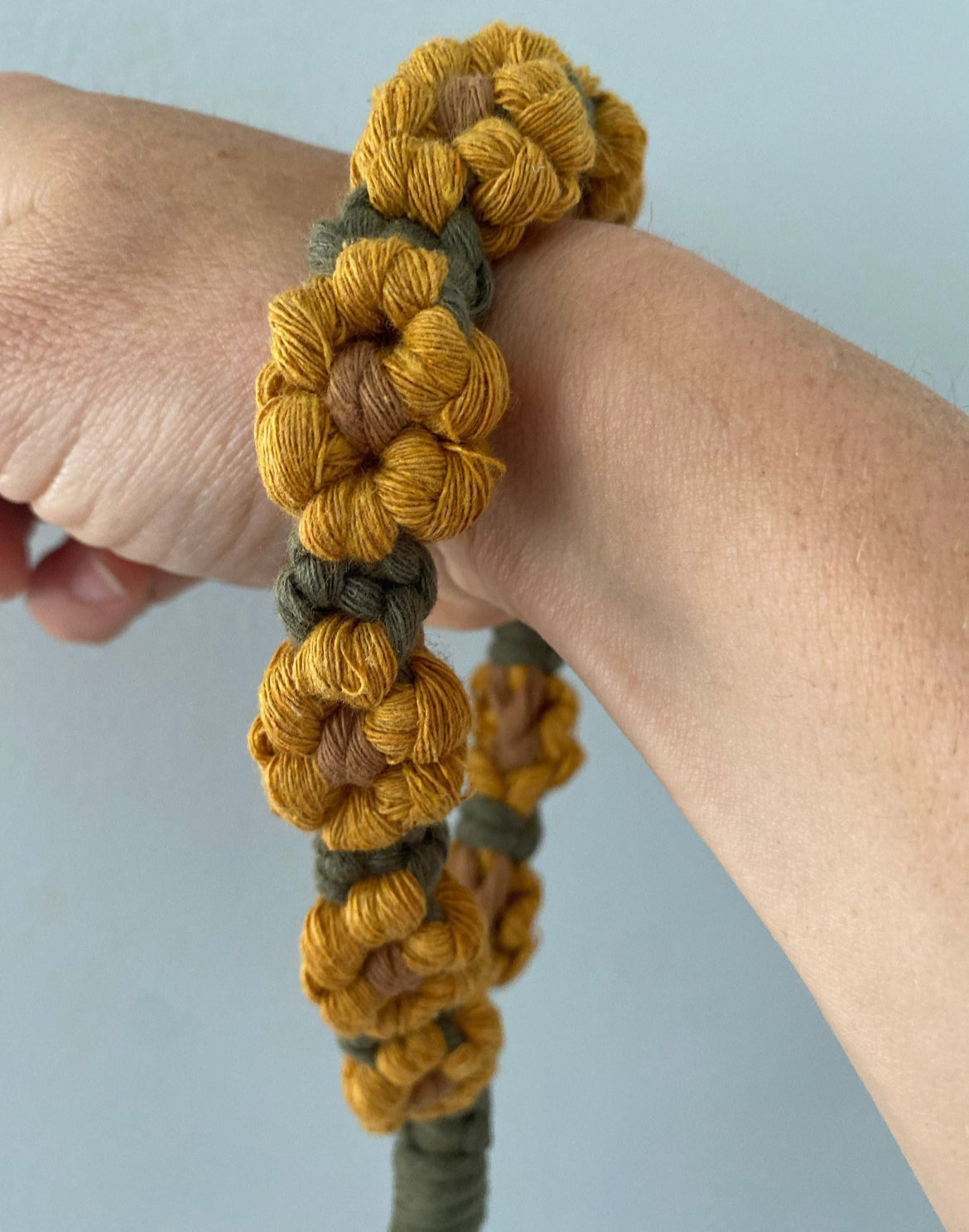 A yellow and green sunflower-themed macramé phone charm wristlet displayed around a person's wrist, with a grey wall in the background. 
