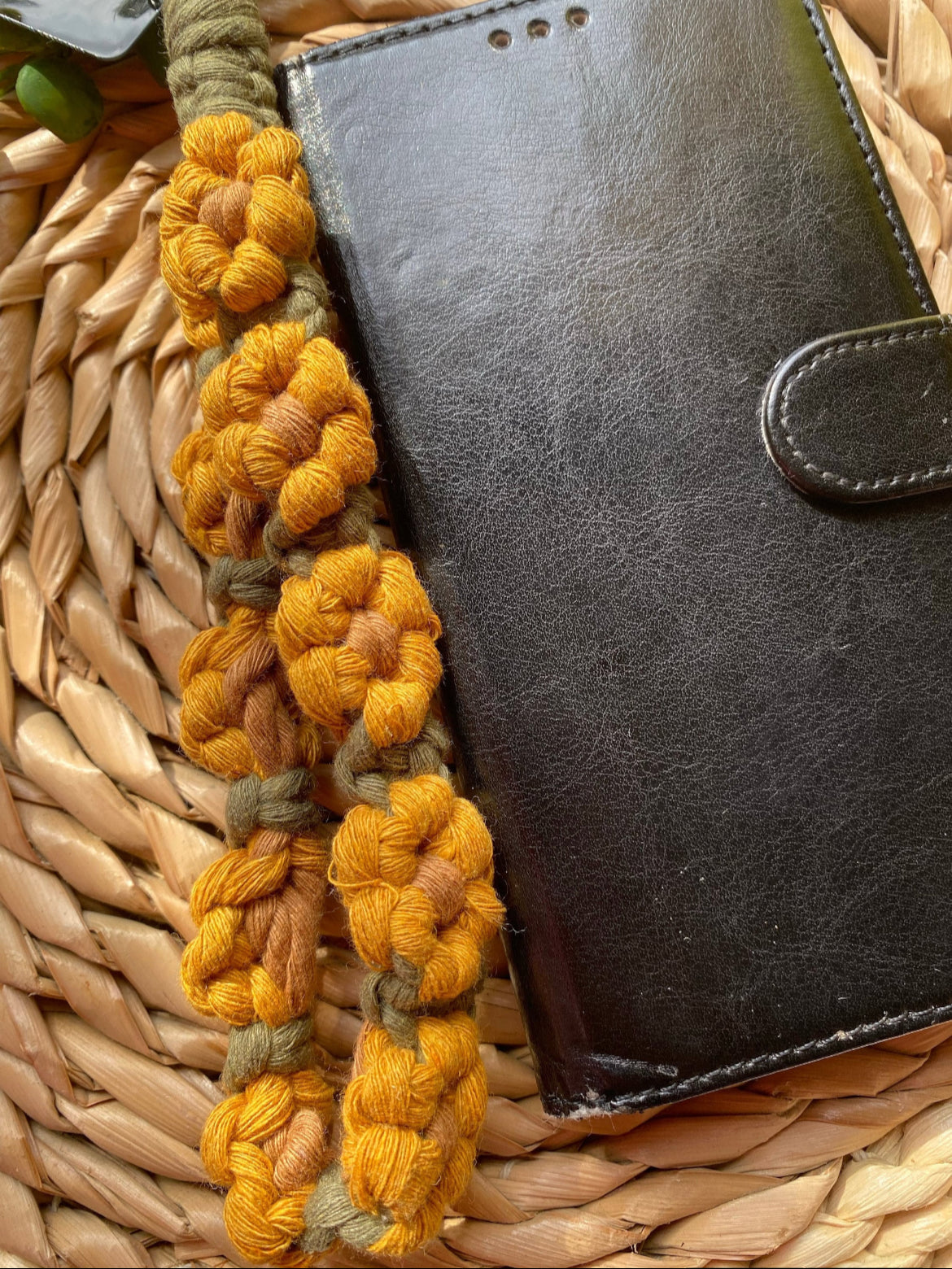 A close up image of a black leather phone case with a yellow and green sunflower-themed macramé phone charm wristlet next to it, on a woven placemat. 