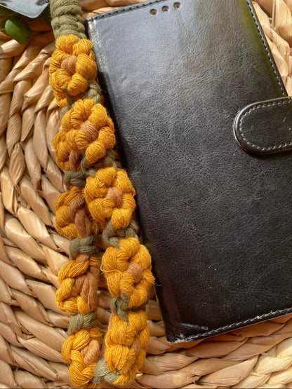 A close up image of a black leather phone case with a yellow and green sunflower-themed macramé phone charm wristlet next to it, on a woven placemat. 