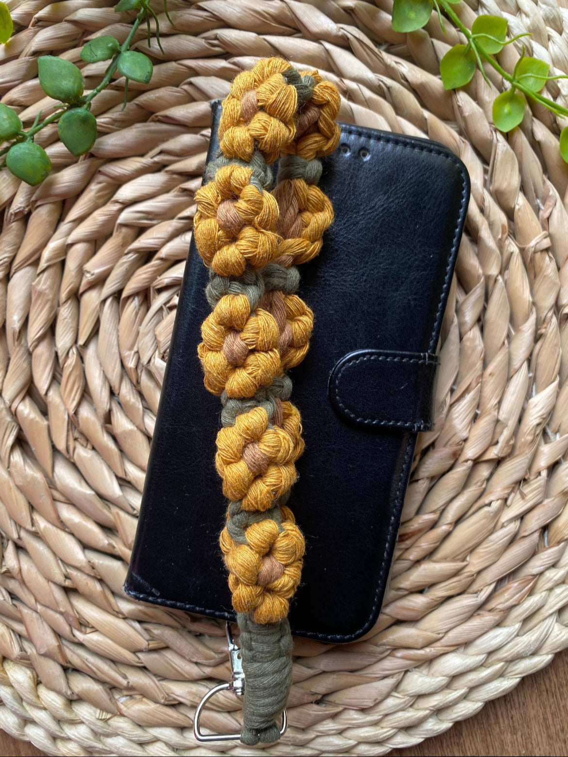 A black leather phone case with a yellow and green sunflower-themed macramé phone charm wristlet placed across it, on a woven mat. 