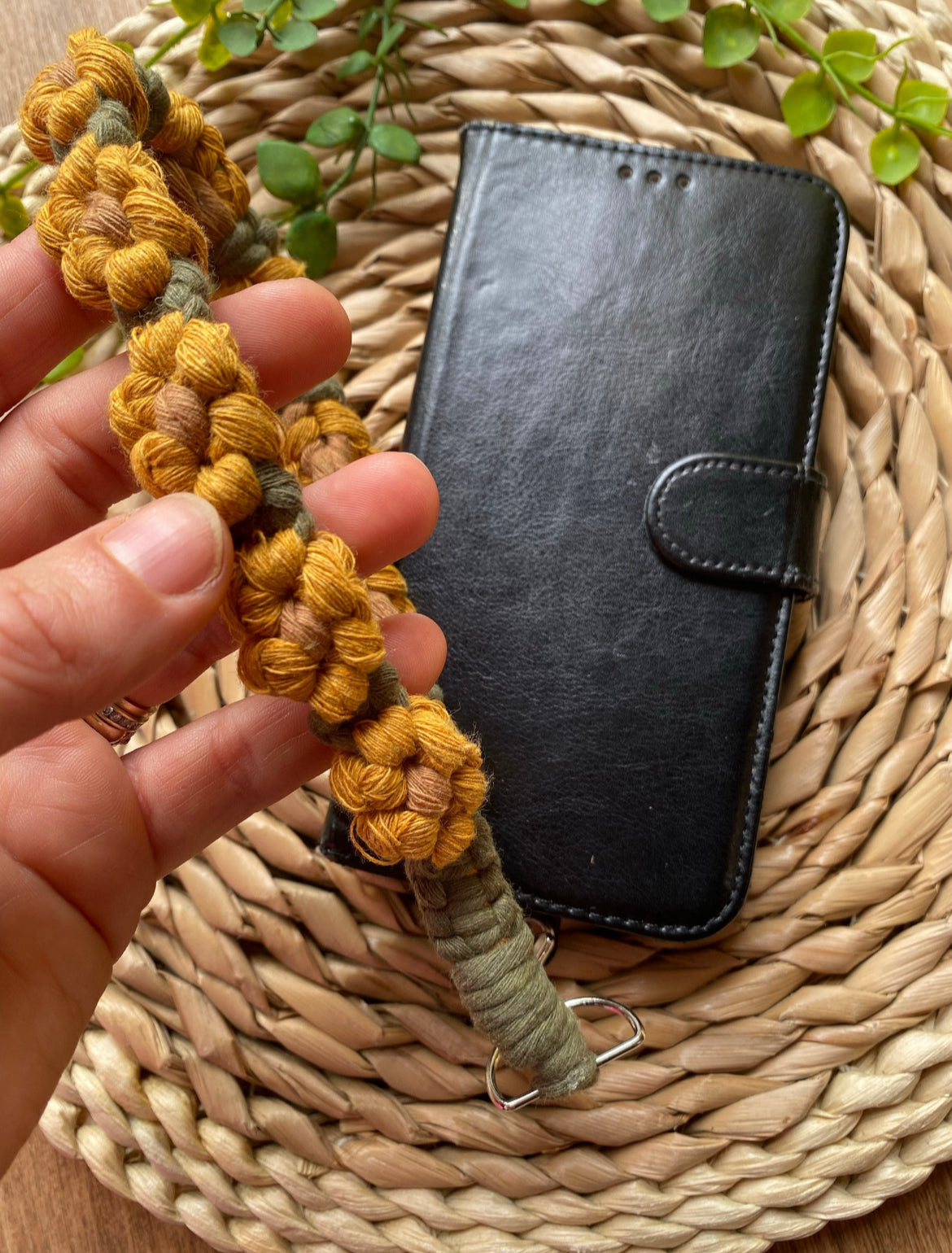 A black leather phone case is placed on a woven mat, a hand holds a yellow and green sunflower-themed macramé phone charm wristlet above it. 