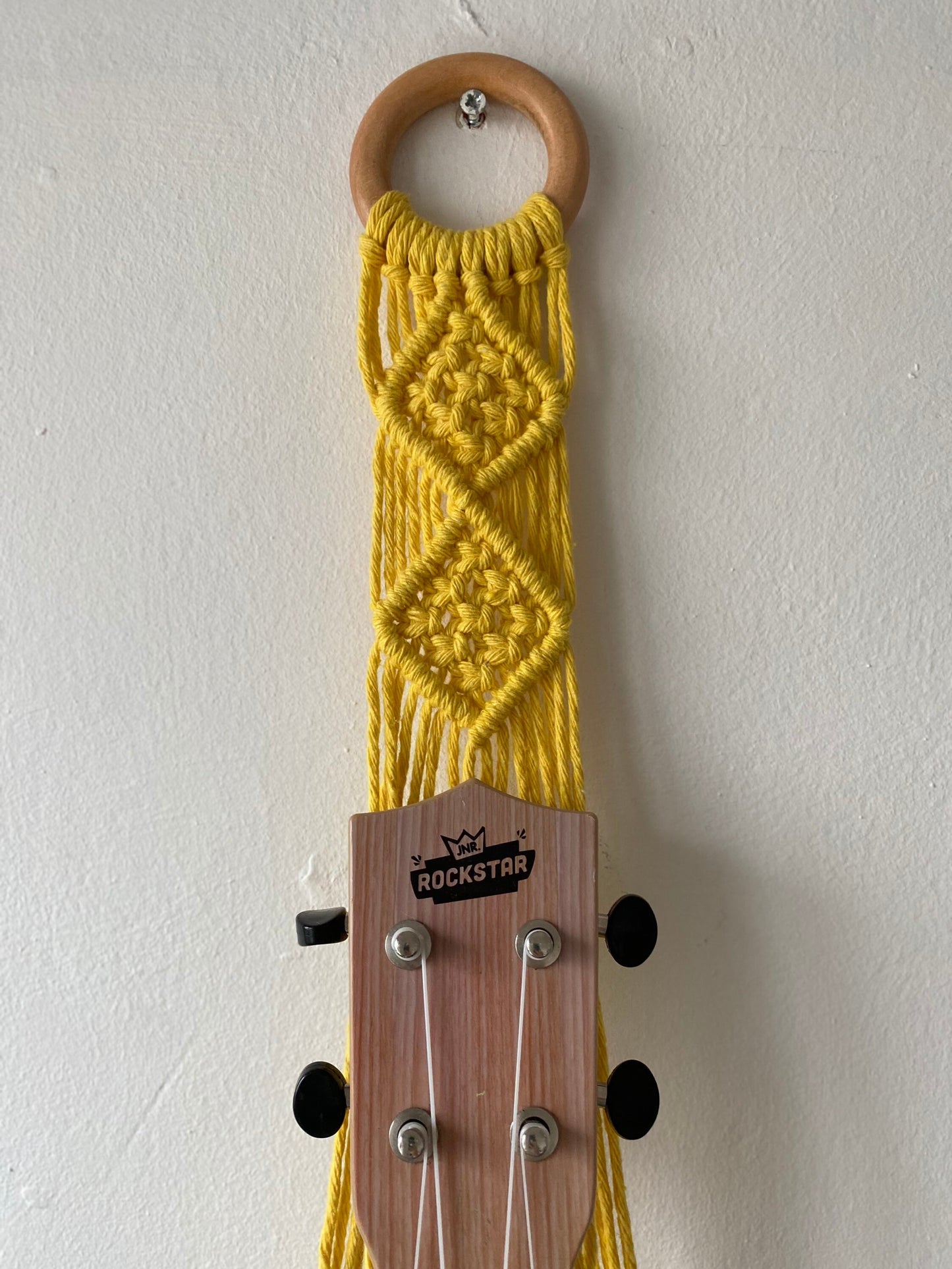 A handmade Ukulele wall mount from Macra-Made-With-Love featuring a wooden ring holding a yellow macrame wall hanging with a diamond pattern, from which a ukulele with a headstock labeled "ROCKSTAR" is suspended. 
