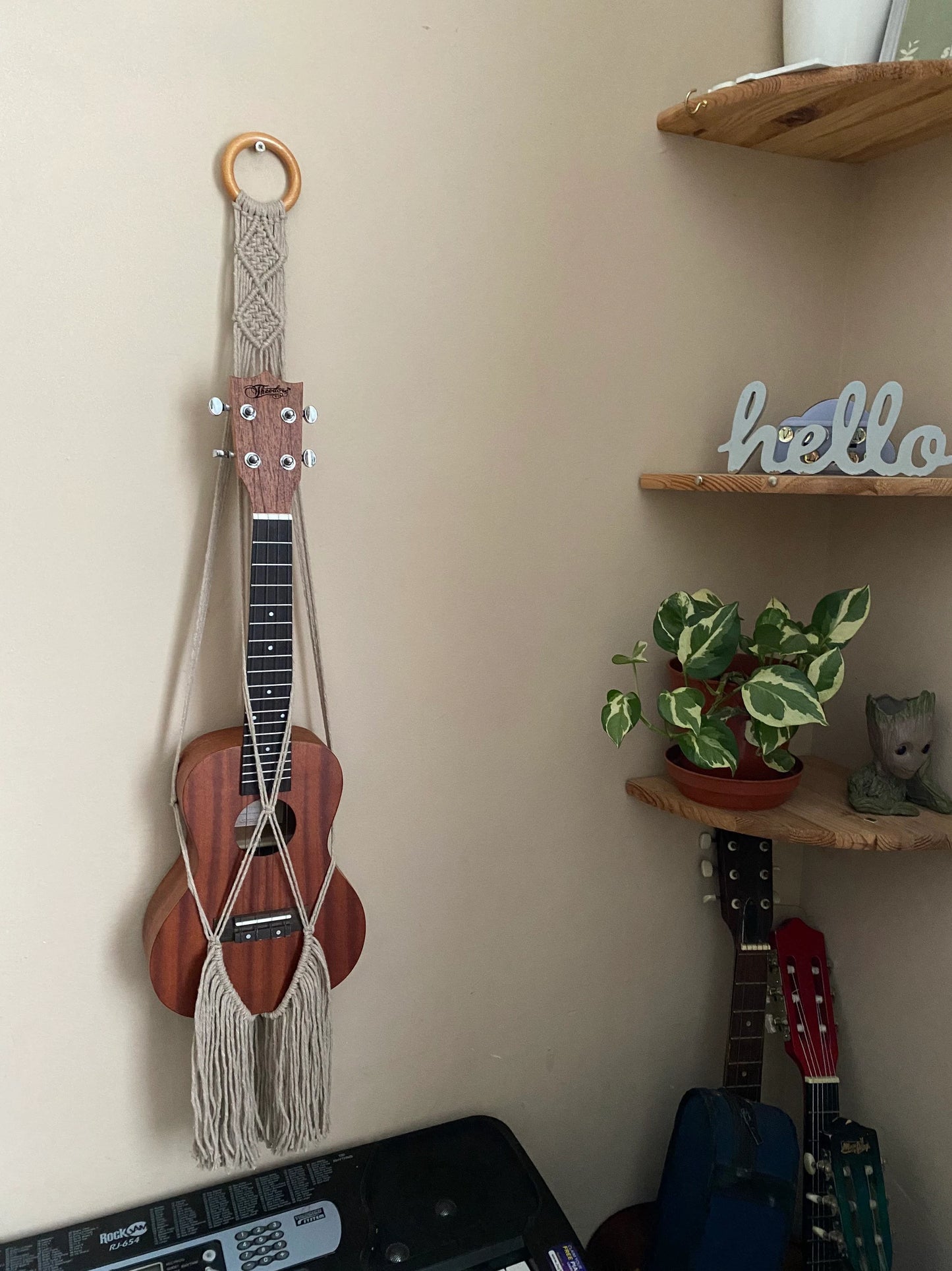 A dark brown ukulele sits inside of a handmade macrame wall hanging ukulele hanger, which is made with taupe coloured recycled cotton, and a repurposed wooden hanging ring. The ukulele hanger also has a long fringe. Three guitars can be seen in the bottom corner of the image under some corner shelves. 