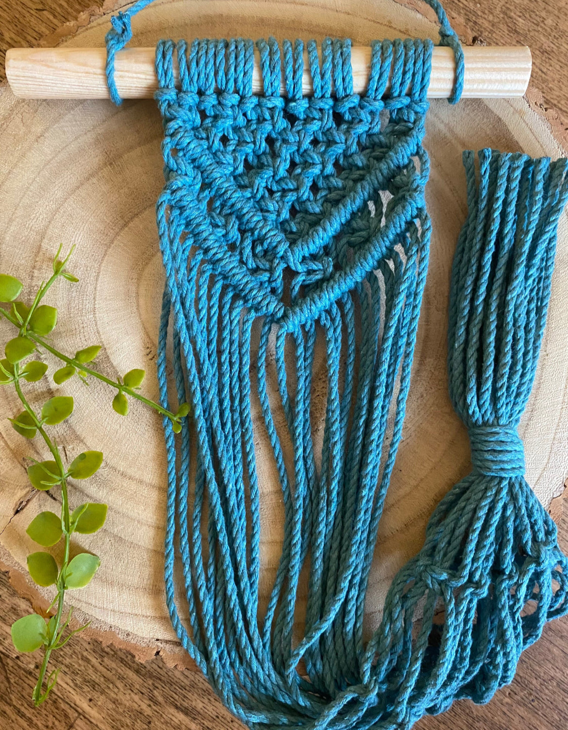 A teal blue macrame wall hanging plant hanger on a wooden dowel, laid flat against a wooden board. 