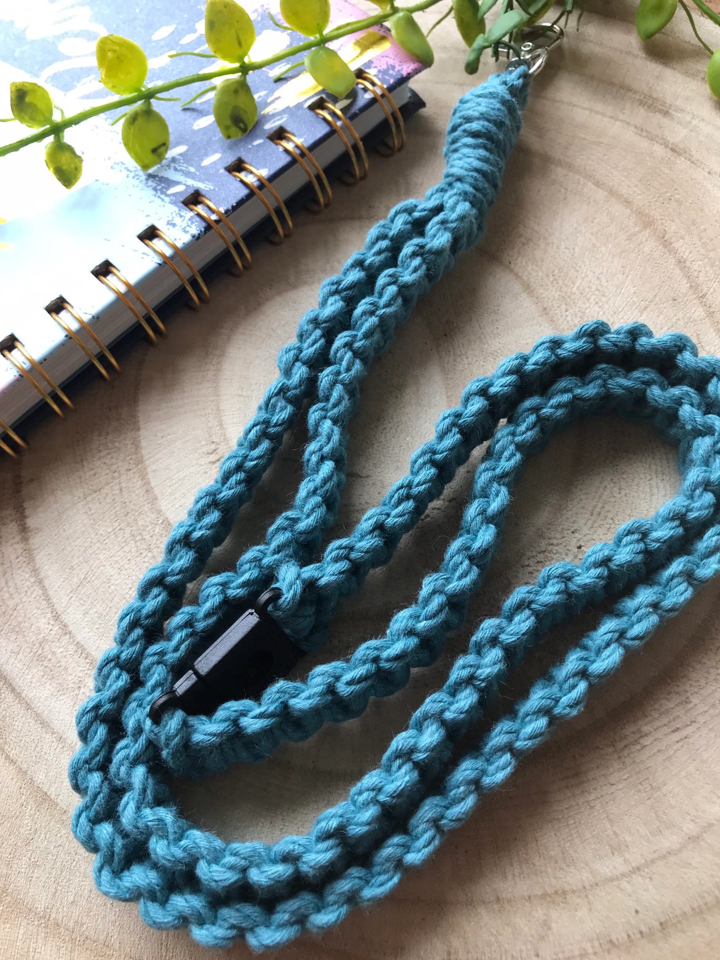 A close-up shot of a teal blue, braided, handmade **Eco friendly lanyard badge holder - 34”** by **Macra-Made-With-Love** with a black plastic clasp lying on a wooden surface. The background includes a spiral-bound notebook with a partial branch of small green leaves resting on it.