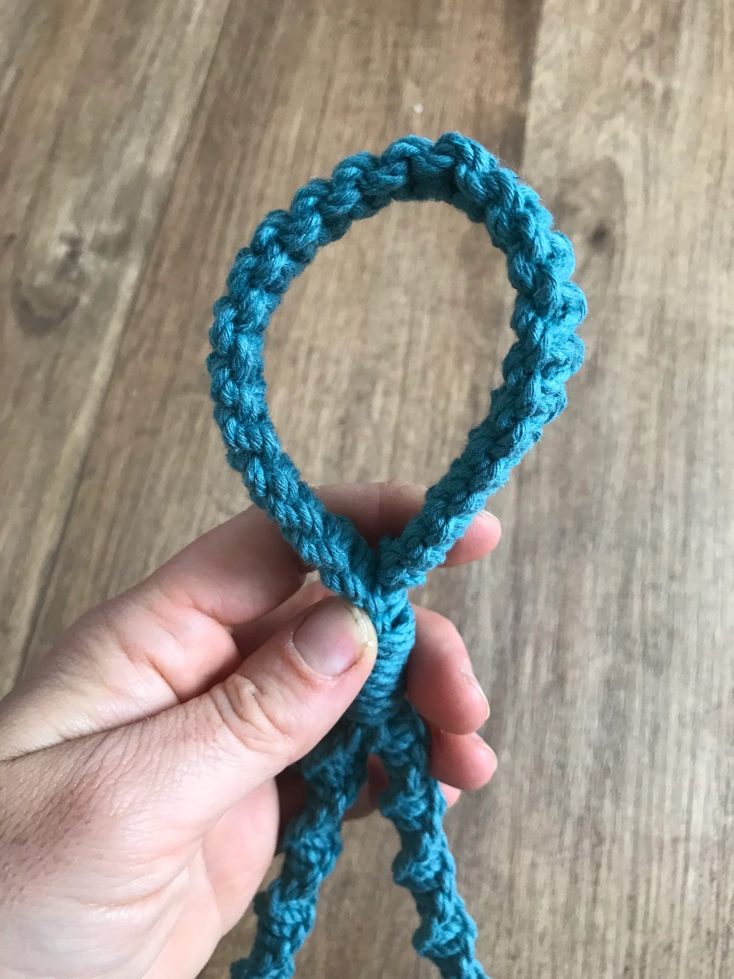 A hand is holding a teal crochet loop against a wooden background. The loop, resembling an eco-friendly decor piece, has a neatly woven pattern. The person's fingers are slightly curled around the end, showcasing the texture and intricacy of the Macra-Made-With-Love Single Toilet Roll Holder.